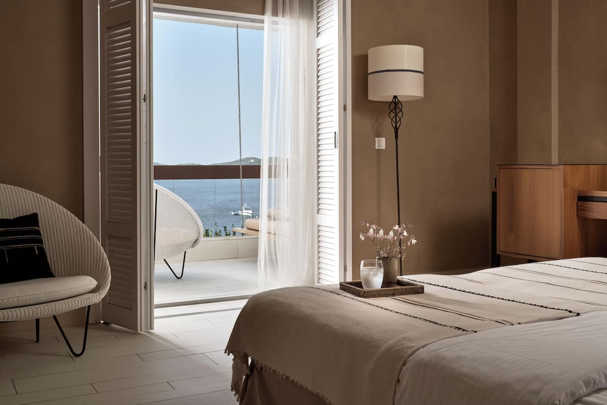A bedroom with a bed, a chair, a lamp, and a view of the sea through a sliding door.