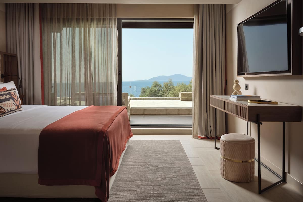 A hotel room with a bed, a desk, a television, and a view of the sea.