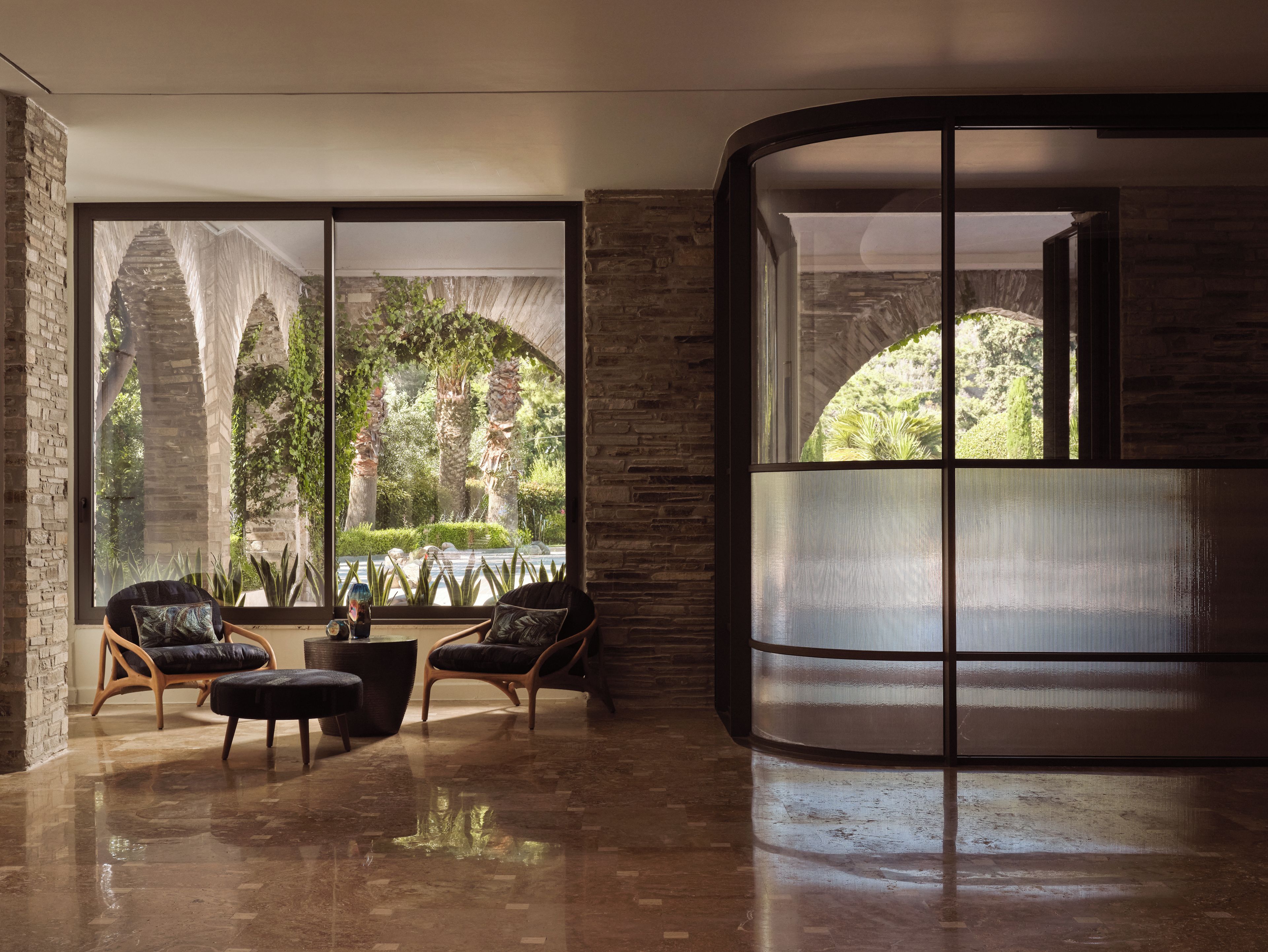 A modern interior with a curved glass partition, a seating area with two chairs and a table, and a view of greenery outside.