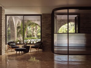 A modern interior with a curved glass partition, a seating area with two chairs and a table, and a view of greenery outside. - Eagles Palace - Small Luxury Hotels of the World (Aristotelis)