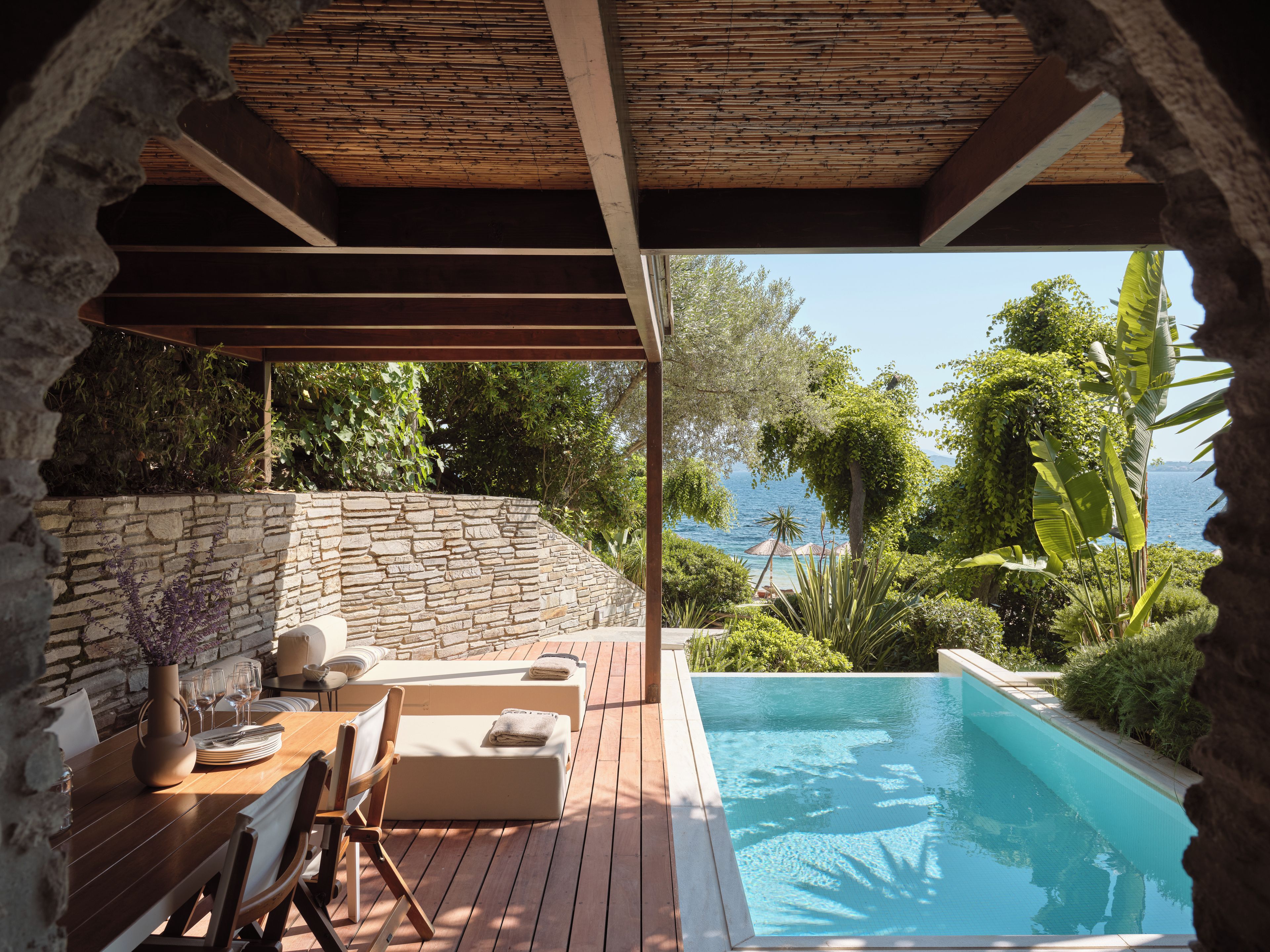 A poolside area with a stone wall, wooden deck, and a view of the sea.