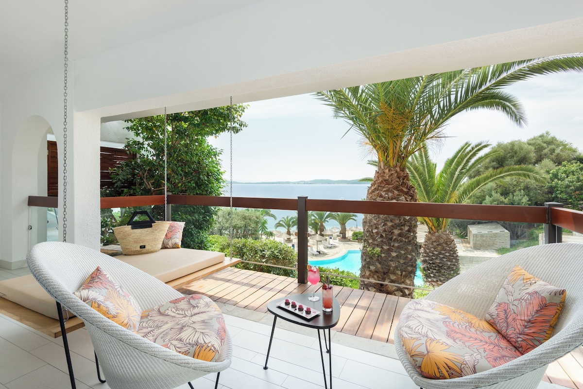 A balcony with wicker chairs, a small table with a drink, and a view of a pool and palm trees.