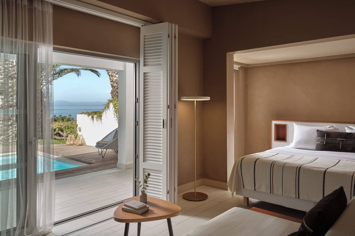 A hotel room with a bed, a bedside table, a lamp, and a view of the pool and ocean.