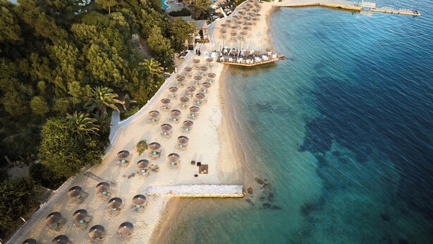 An aerial view of a beach resort with rows of sun loungers and umbrellas, a boat dock, and a clear blue sea.