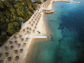 An aerial view of a beach resort with rows of sun loungers and umbrellas, a boat dock, and a clear blue sea. - Eagles Palace - Small Luxury Hotels of the World (Aristotelis)