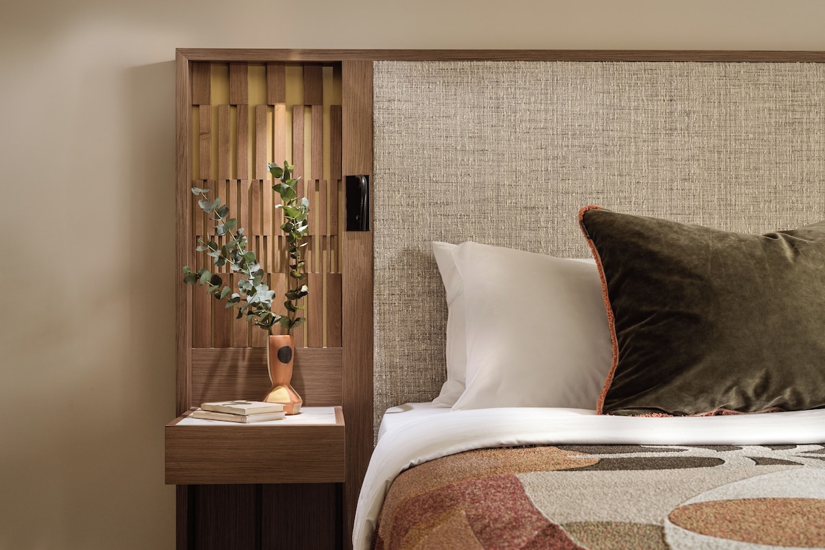 A neatly arranged bedroom with a wooden nightstand, a vase with a plant, a bed with white and brown bedding, and a textured headboard.