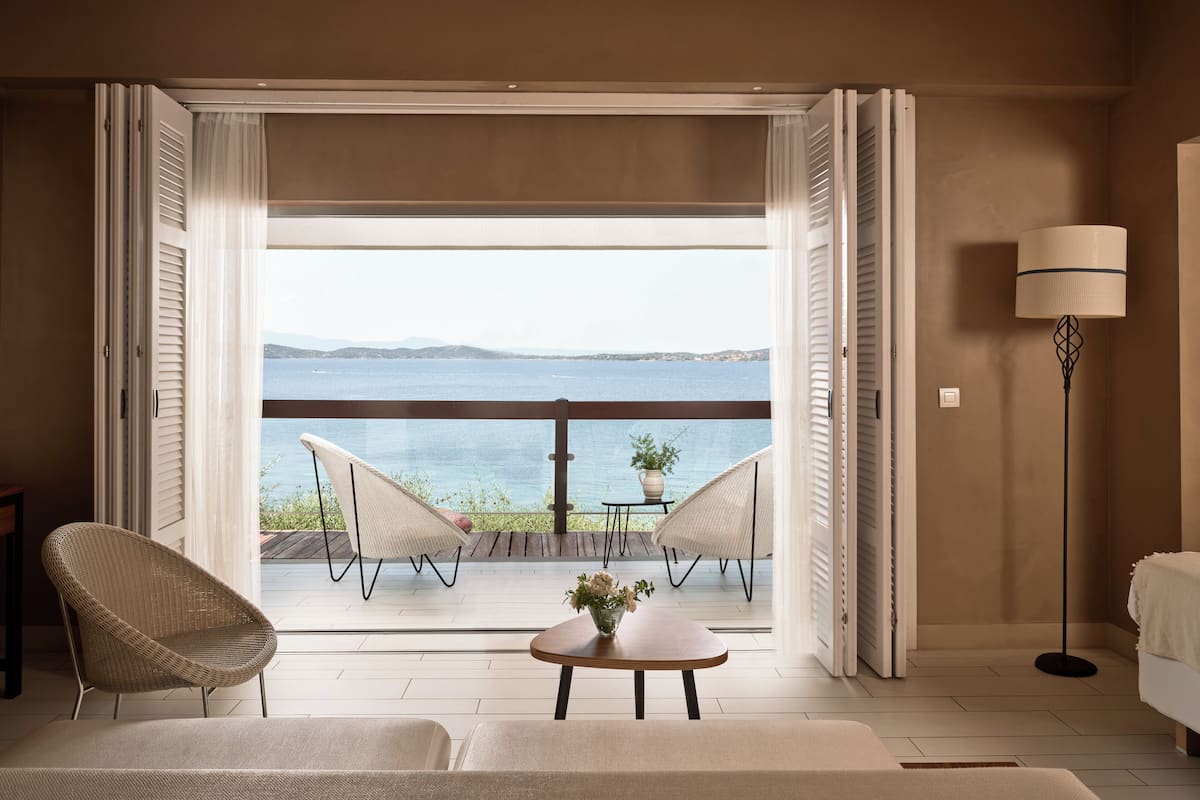 A room with a balcony overlooking a body of water, furnished with a sofa, chairs, and a small table with a vase.
