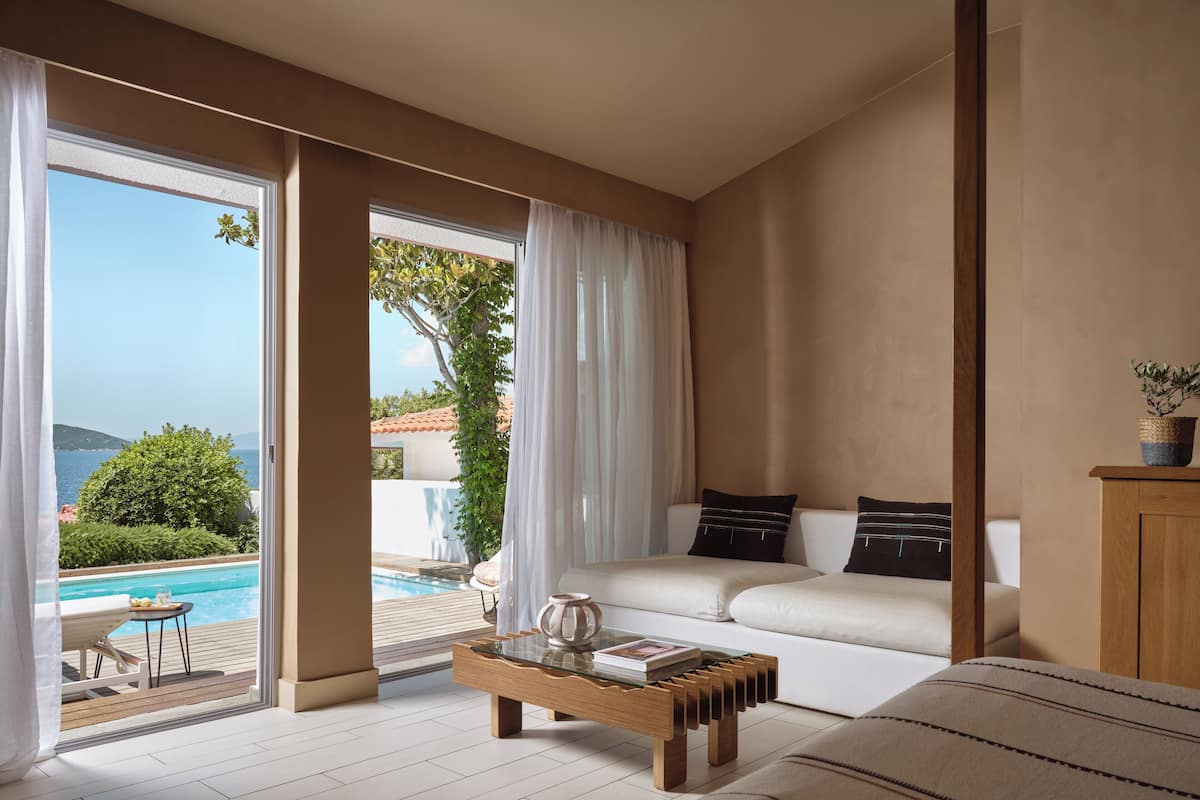 A modern bedroom with a large window, a wooden coffee table, a sofa, and a view of a pool and greenery.