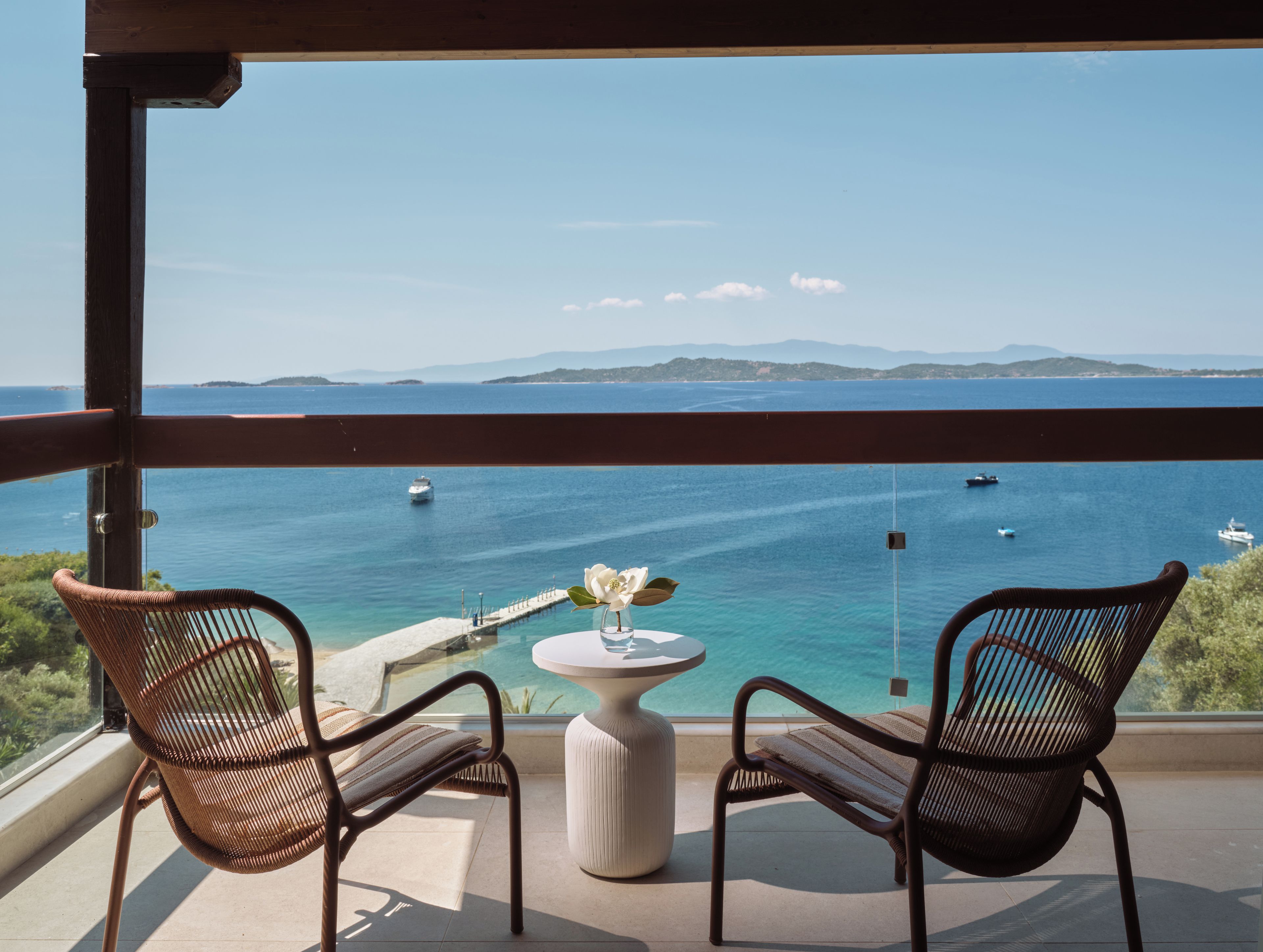 A balcony with two wicker chairs and a small table with a flower vase, overlooking a clear blue sea with boats and distant land.