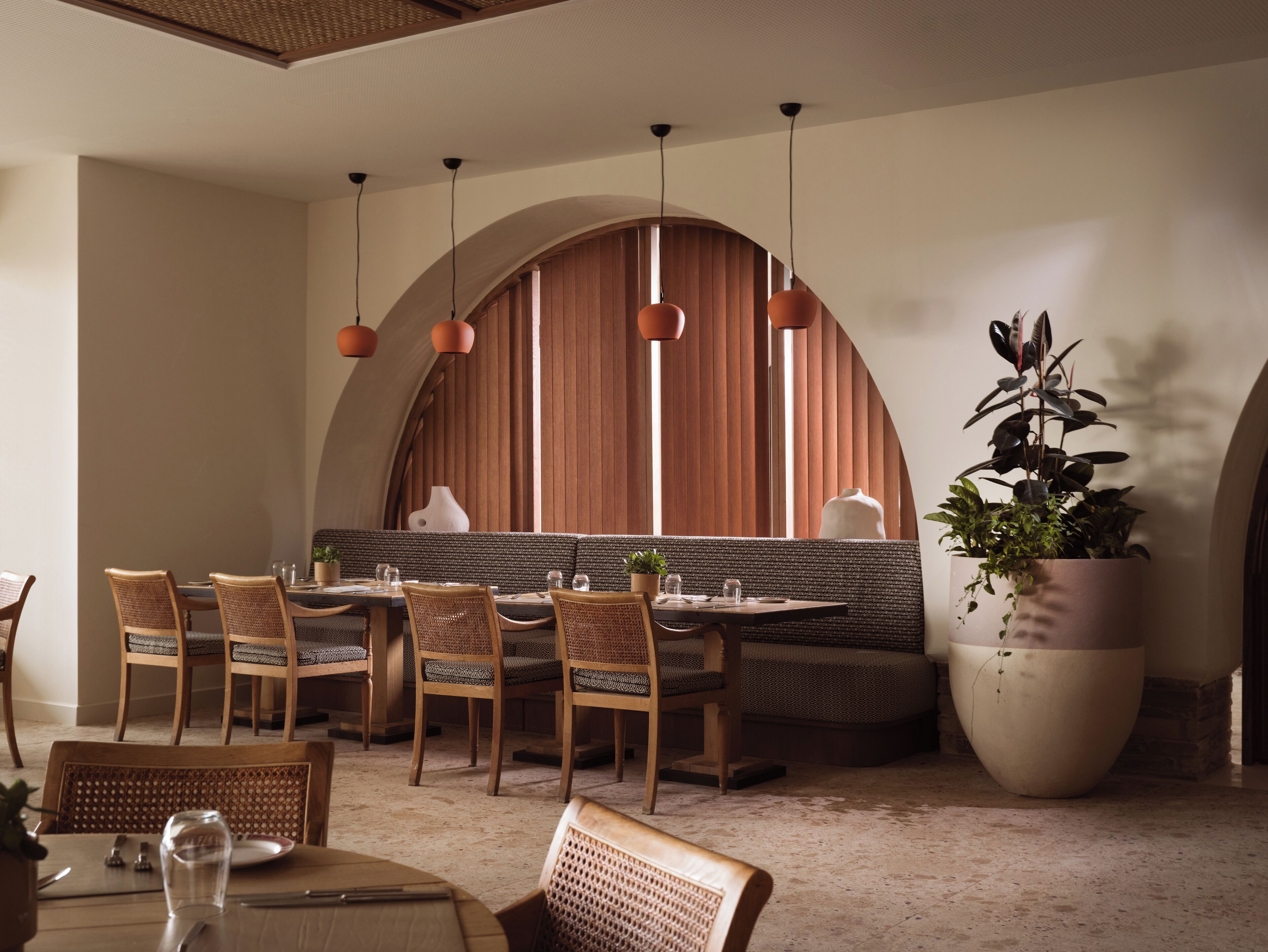 Un comedor con mesas y sillas de madera, una planta grande en maceta y un diseño de pared en arco original.