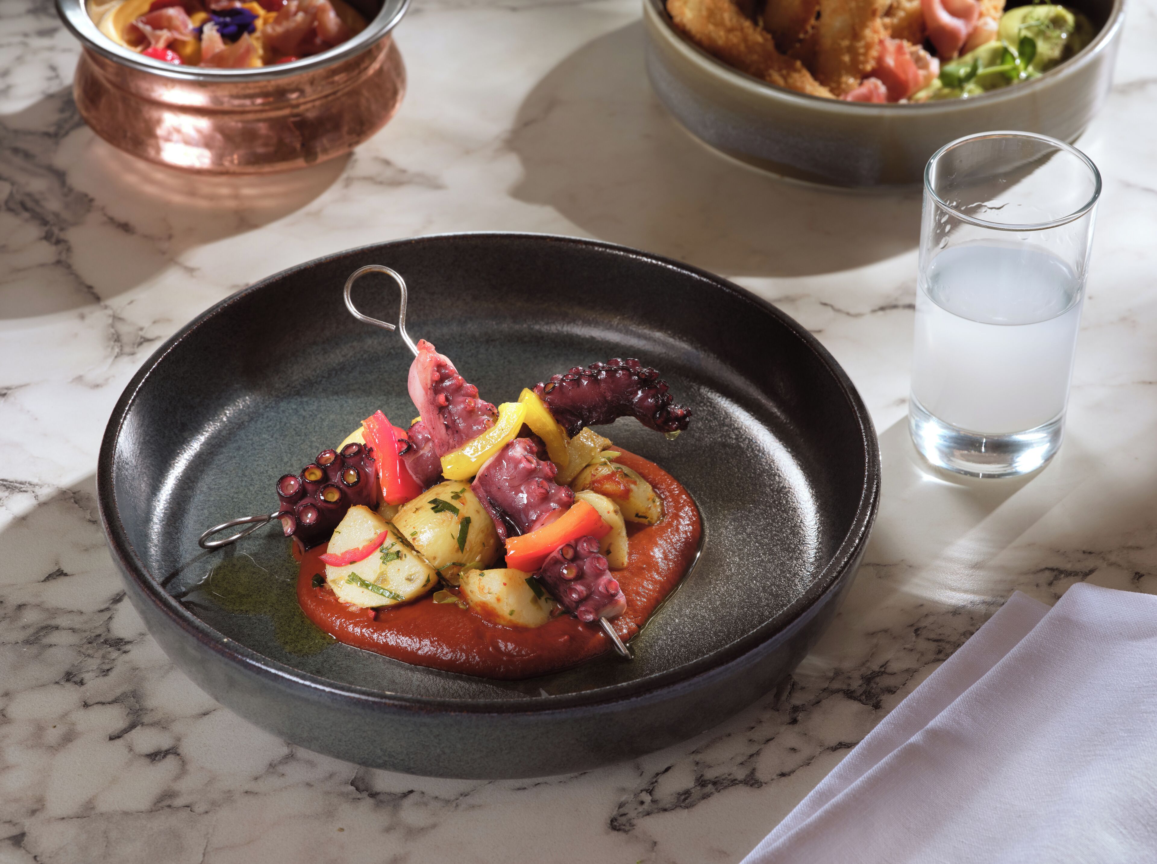 Un plat contenant des poulpes et des légumes, servi sur une assiette noire, avec un verre d’eau et un bol en cuivre en arrière-plan.