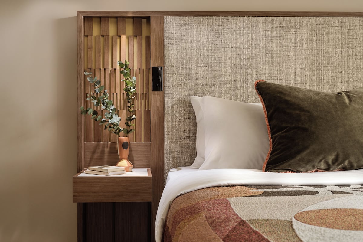 A neatly arranged bedroom with a wooden nightstand, a vase with a plant, a bed with white and brown bedding, and a textured headboard.