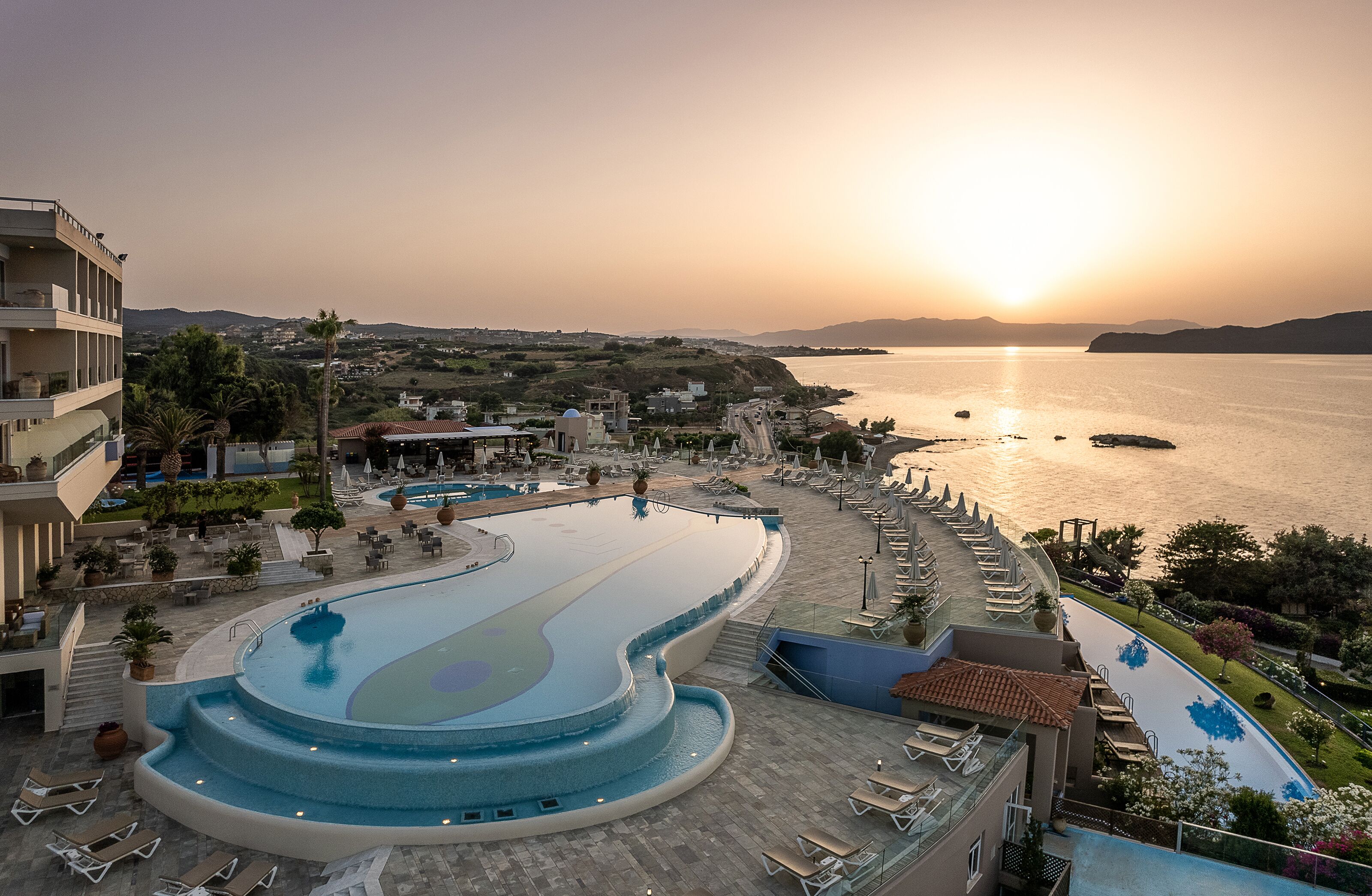 A resort pool with a curved design, surrounded by lounge chairs and a view of the sea at sunset.