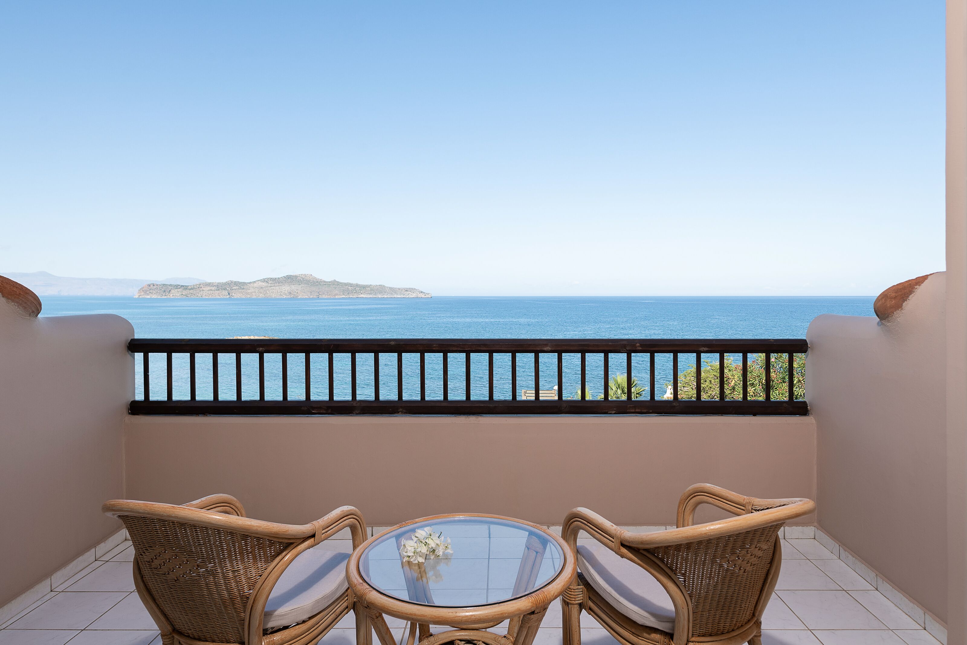 A balcony with wicker furniture and a glass table, overlooking a clear blue sea and a distant island.