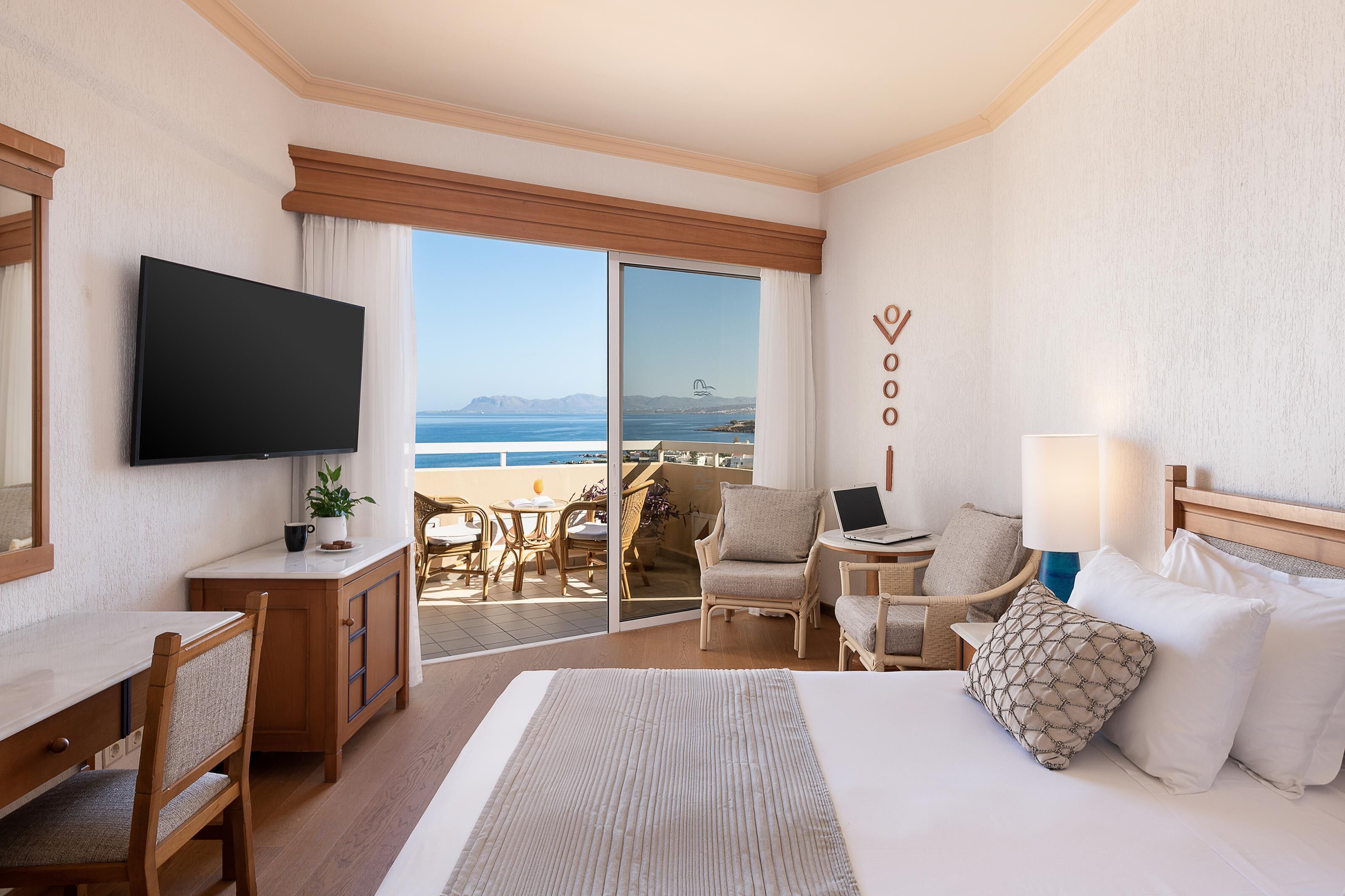 A hotel room with a bed, a desk, a TV, a laptop, and a view of the ocean.