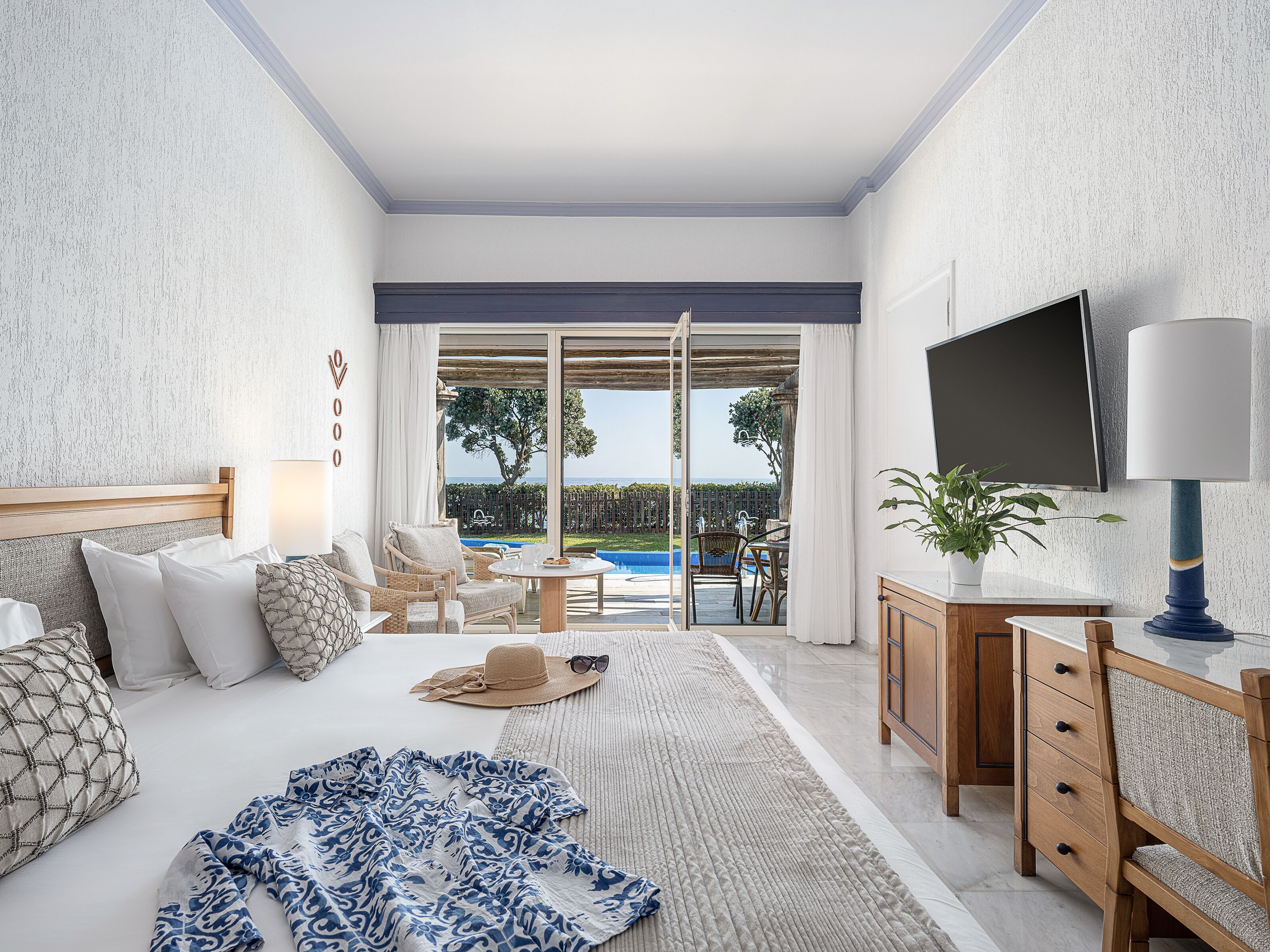 A bedroom with a large bed, a TV, a desk, and a view of a pool and garden.