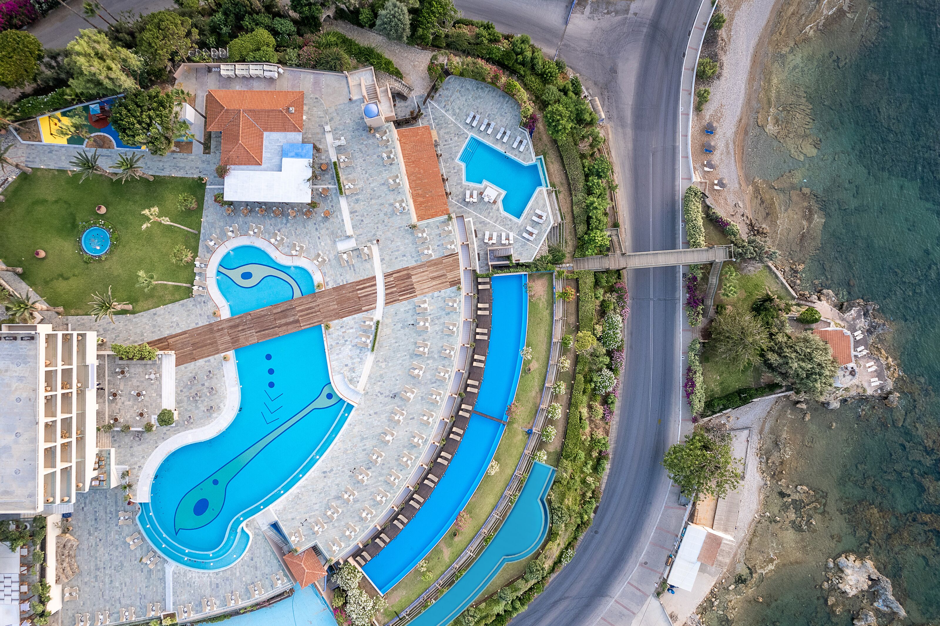 An aerial view of a resort with multiple swimming pools, a bridge, and a coastal road.