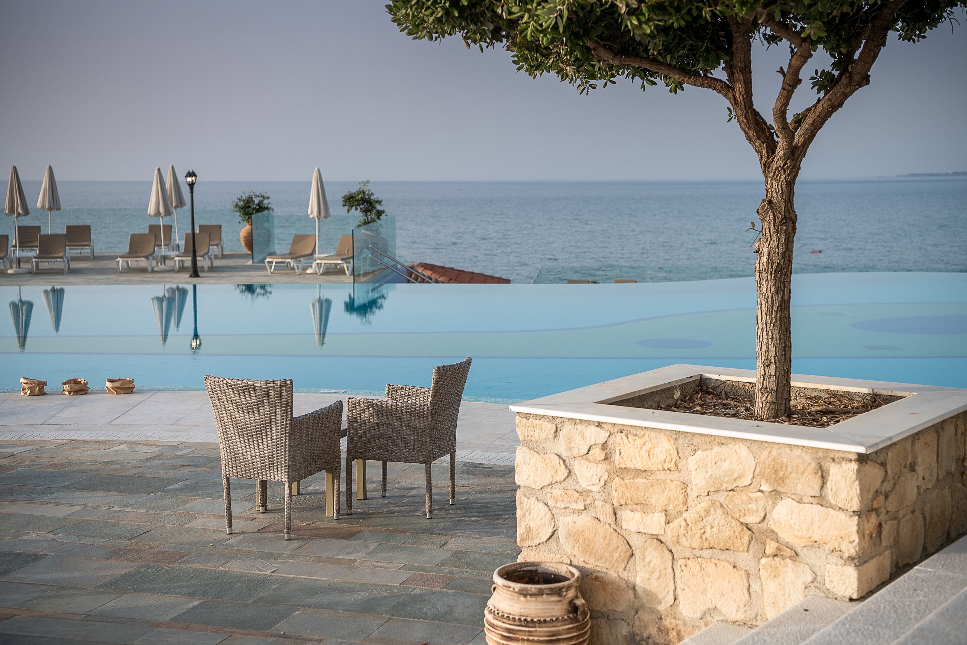 Un área junto a la piscina con sillas de descanso y sombrillas, una jardinera de piedra con un árbol, y vista al mar.