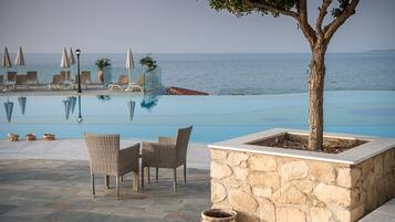 Un área junto a la piscina con sillas de descanso y sombrillas, una jardinera de piedra con un árbol, y vista al mar.
