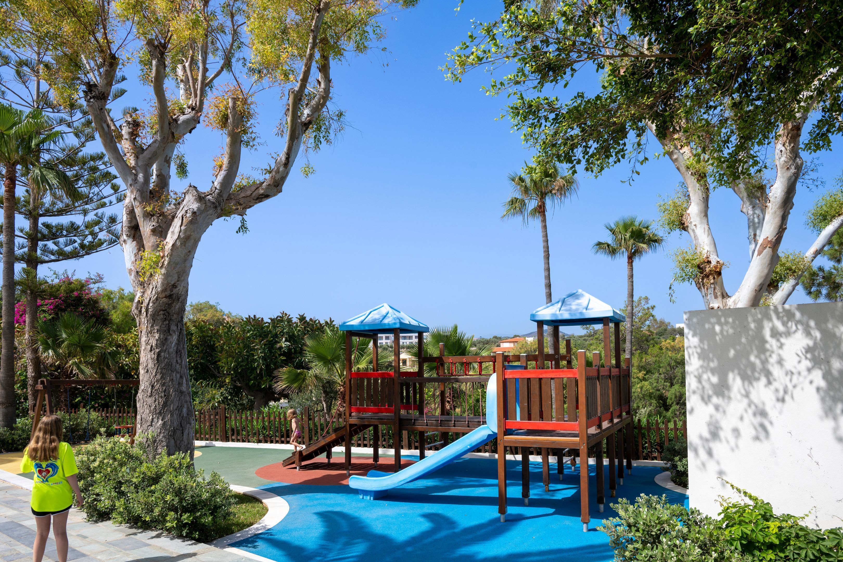 A playground with a slide and climbing structures, surrounded by trees and a clear sky.
