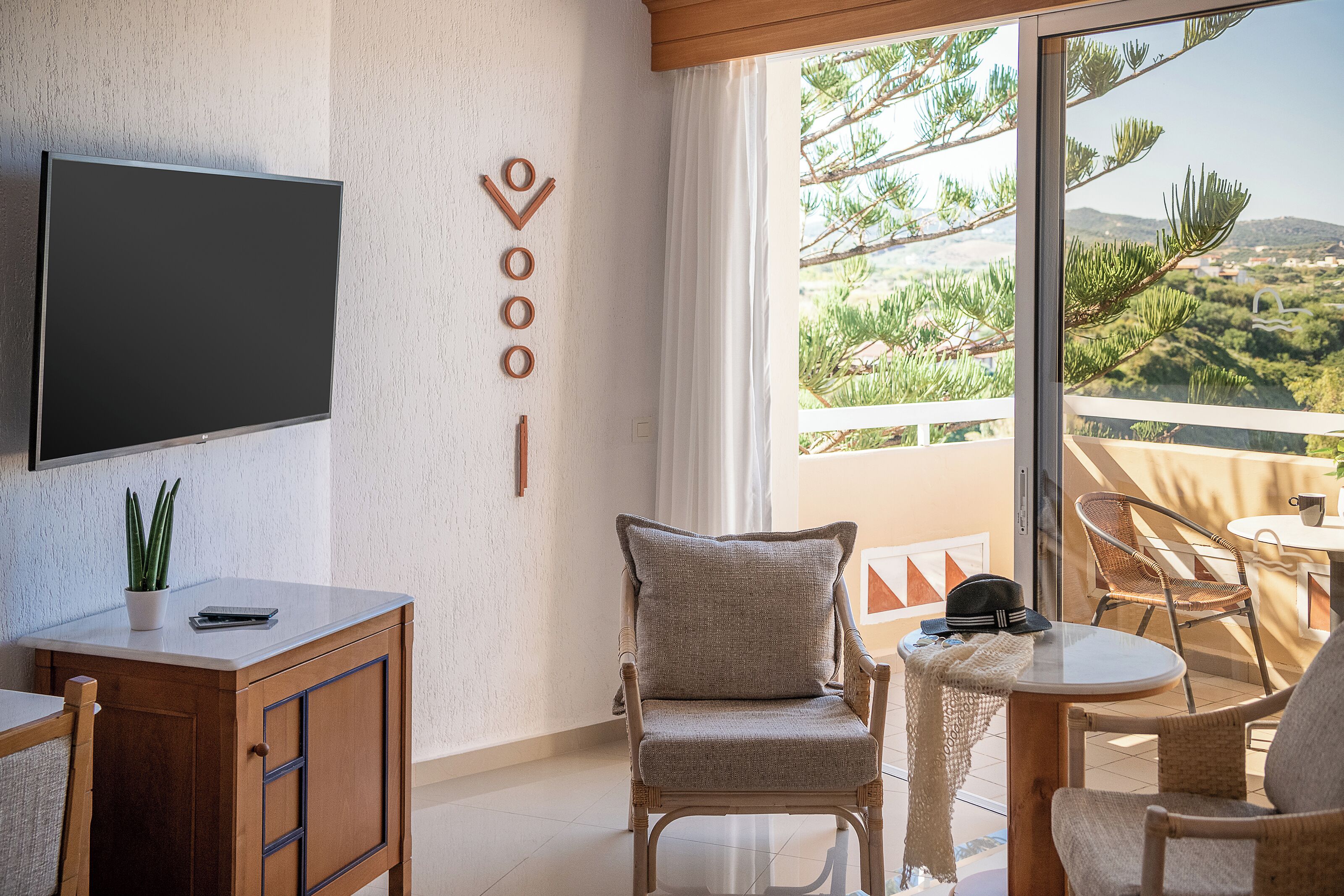 Uma sala de estar moderna com televisão de tela plana, uma poltrona confortável, uma mesinha e uma varanda com vista para vegetação e colinas.