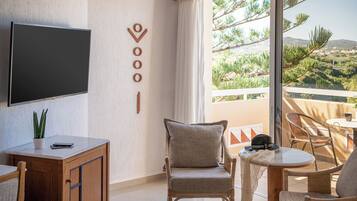 Uma sala de estar moderna com televisão de tela plana, uma poltrona confortável, uma mesinha e uma varanda com vista para vegetação e colinas.