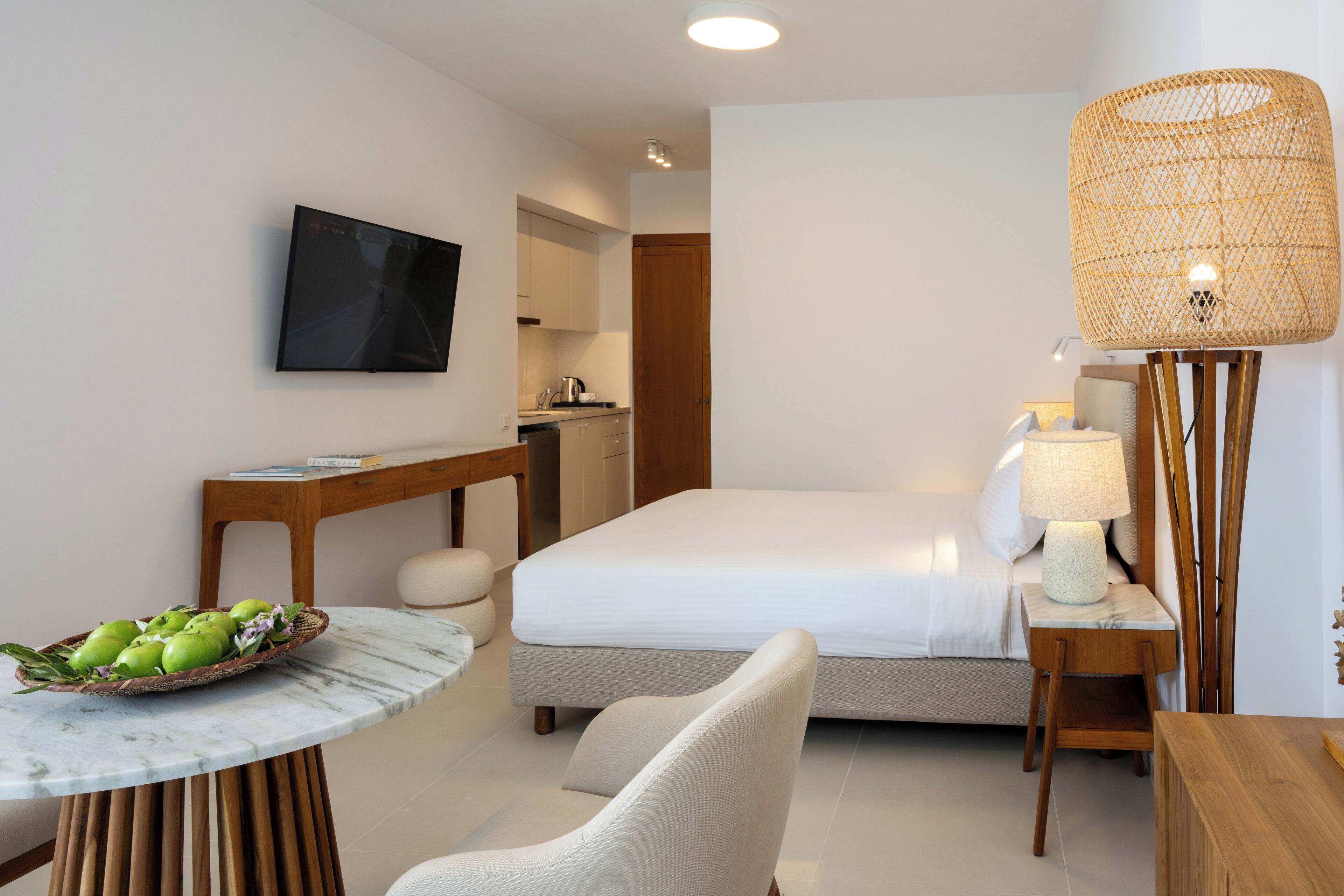 Una habitación de hotel moderna con una cama, un escritorio con una computadora, una mesa de comedor con manzanas, un televisor y una lámpara.