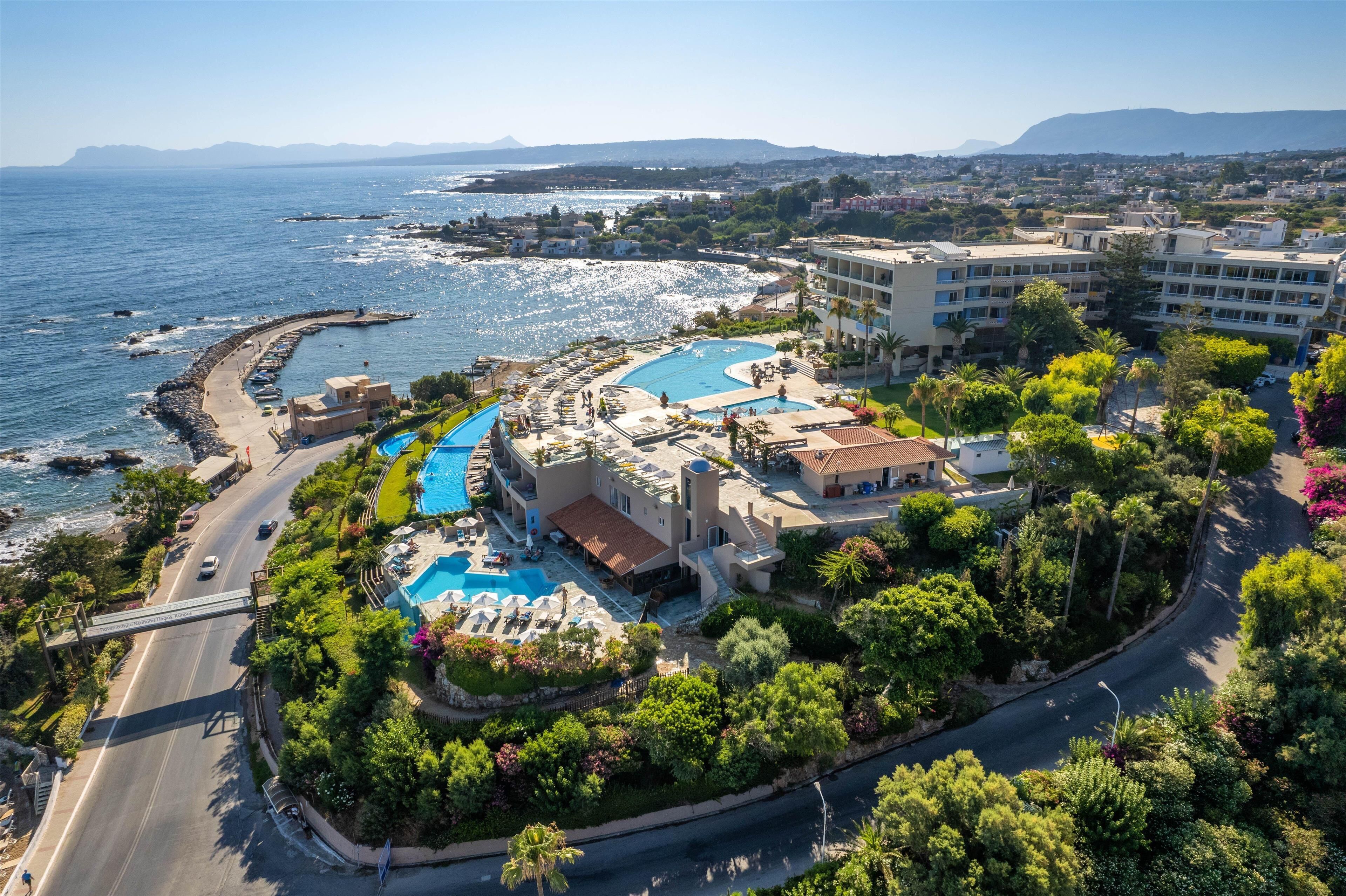 An aerial view of a coastal resort with a swimming pool, a road, and surrounding greenery.