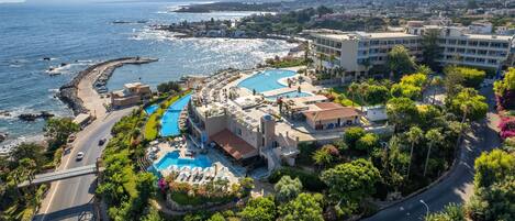 An aerial view of a coastal resort with a swimming pool, a road, and surrounding greenery.