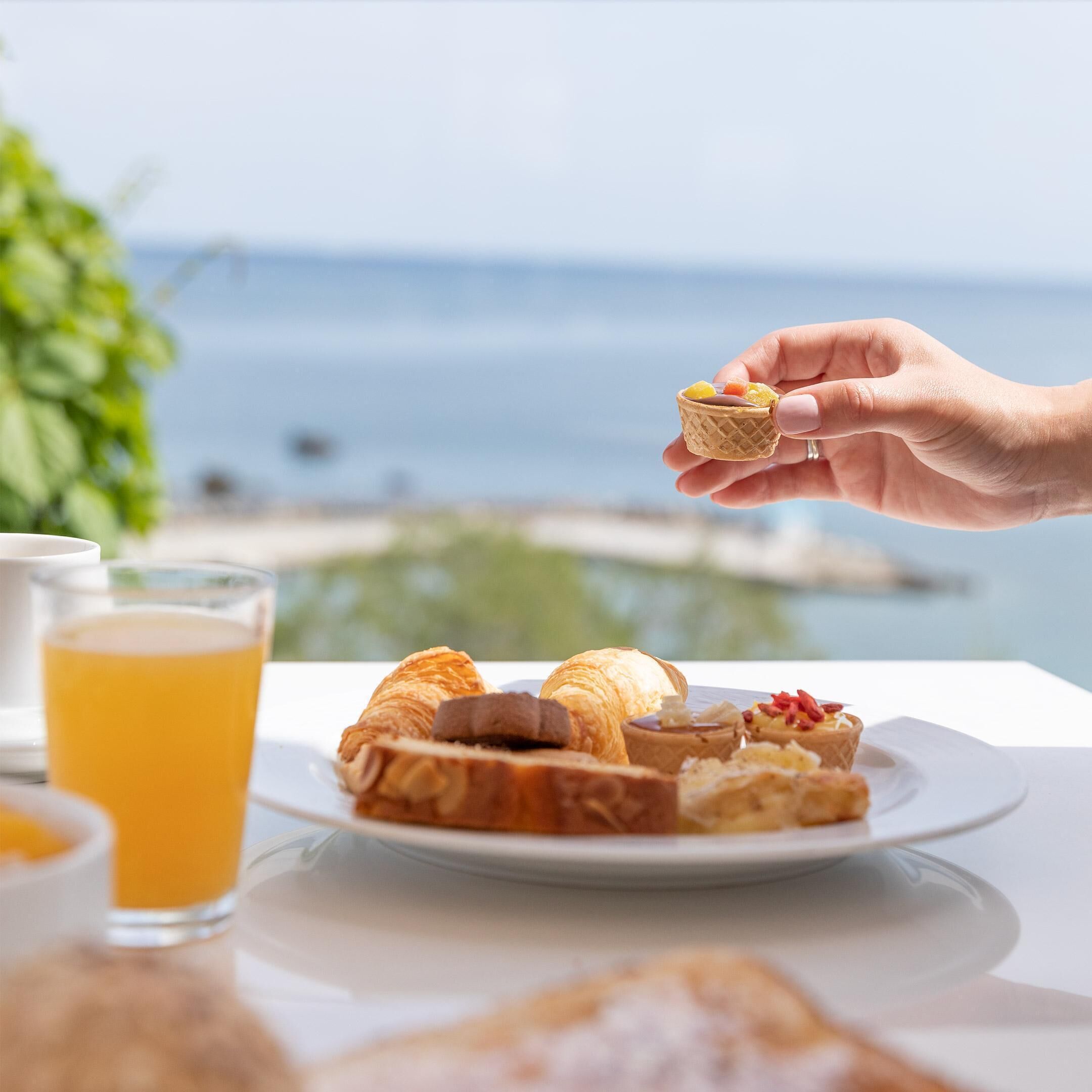 Une table avec une assiette de pâtisseries et un verre de jus d’orange, donnant sur une plage.
