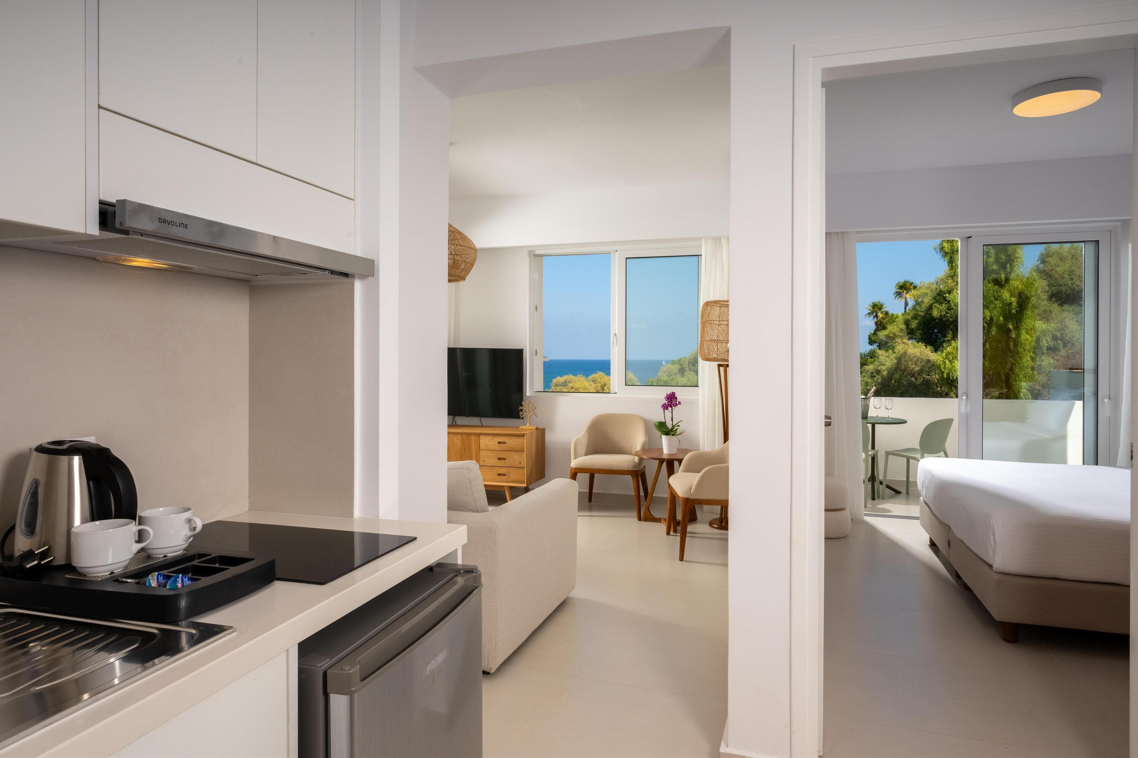 A modern hotel room with a kitchenette, a dining area, a sofa, a bed, and a balcony with a view of the sea.