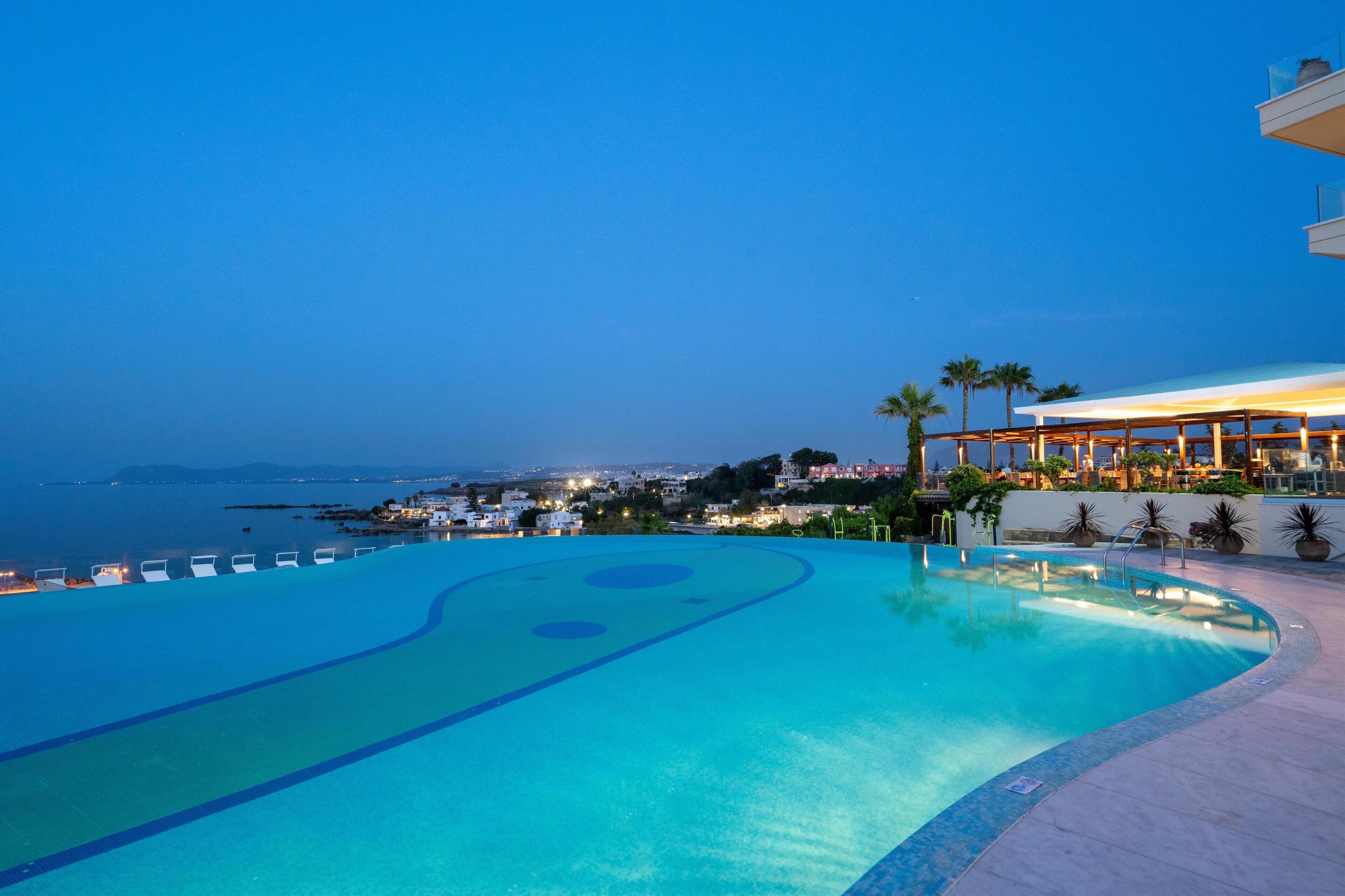 A pool with a clear view of the sea and distant cityscape, surrounded by palm trees and a building with a covered area.
