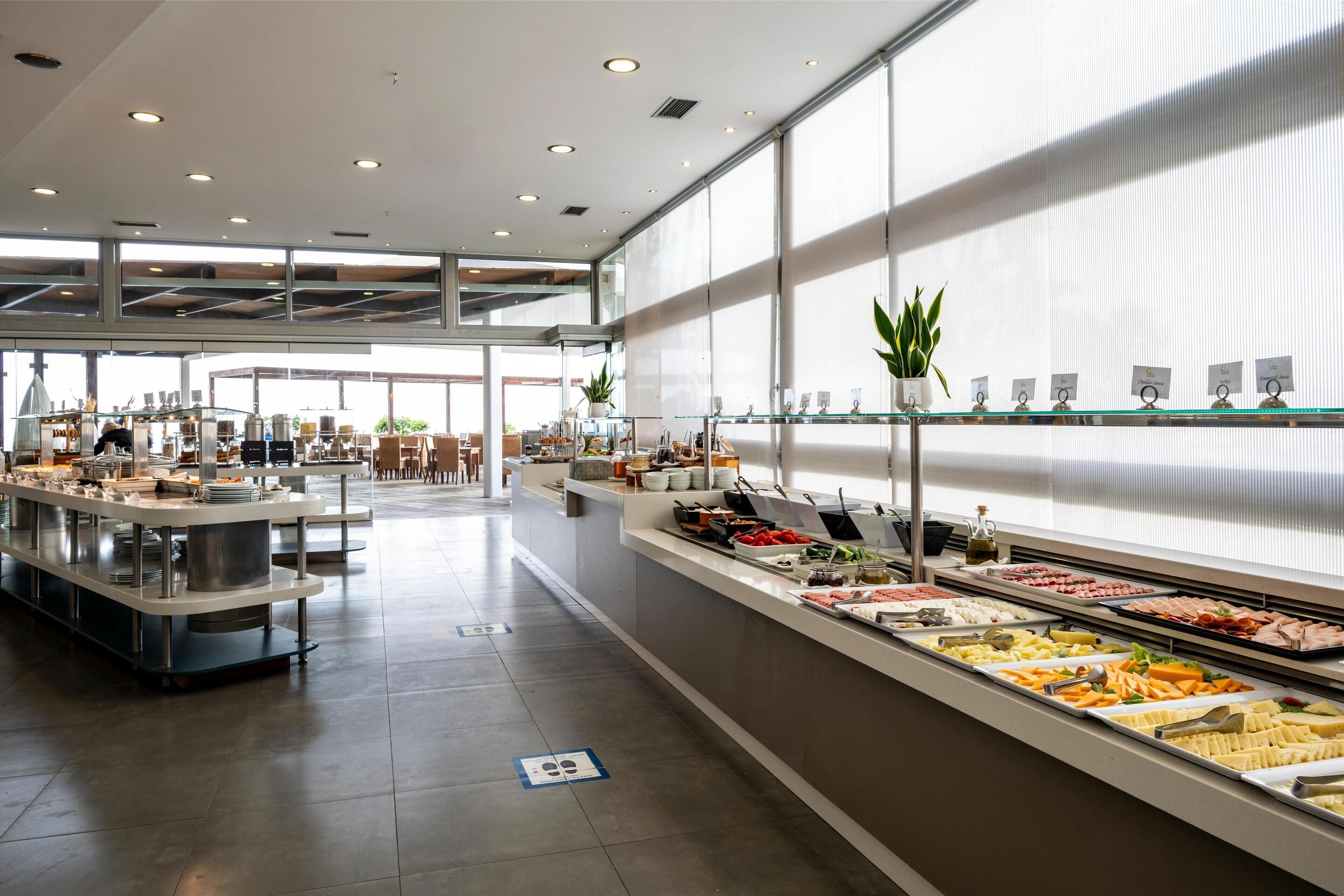 Zona de bufé con diversas opciones de comida, una zona de estar y grandes ventanales que dejan entrar luz natural.