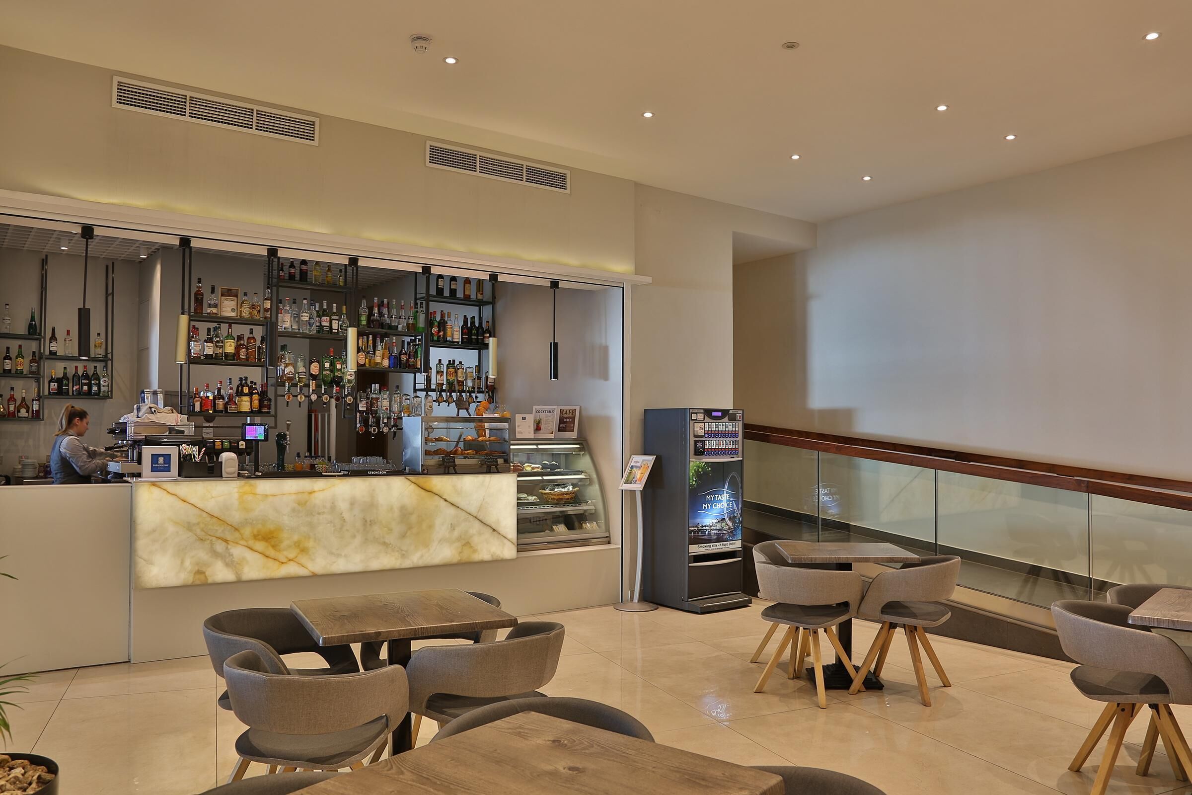 A modern bar area with marble countertops, shelves of liquor bottles, and a digital menu kiosk. There are several tables and chairs for patrons.