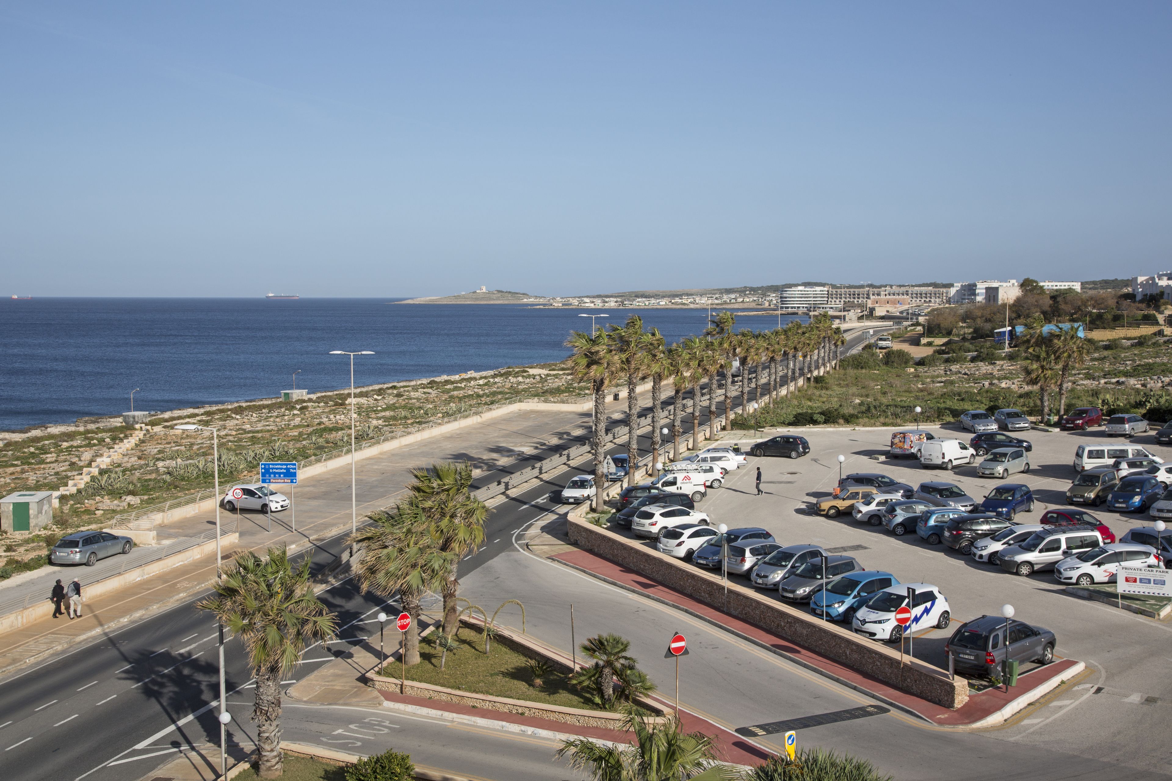 Un estacionamiento frente al mar, una carretera bordeada de palmeras y cielo despejado.