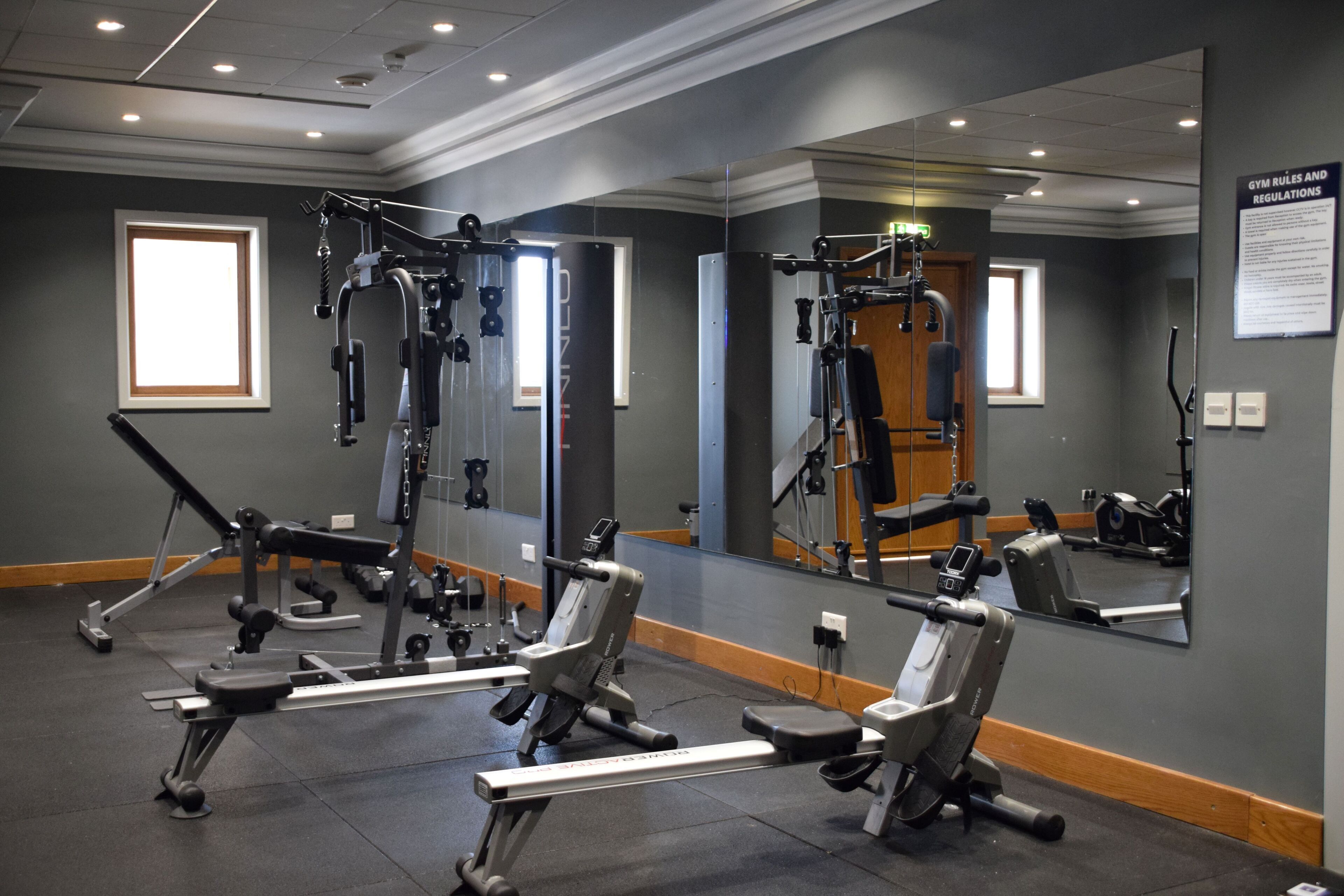 Een fitnessruimte met diverse fitnessapparaten, een grote spiegel, en een bord met regels en voorschriften.
