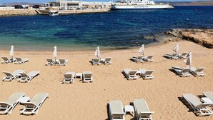Een strand met witte ligbedden en parasols, een pier met een grote witte boot, en een helderblauwe lucht.