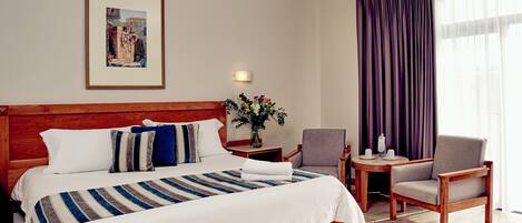 Habitación de hotel con una cama grande, cabecera de madera, un cuadro enmarcado en la pared, una mesita con un florero y una silla, además de una ventana con cortinas moradas.