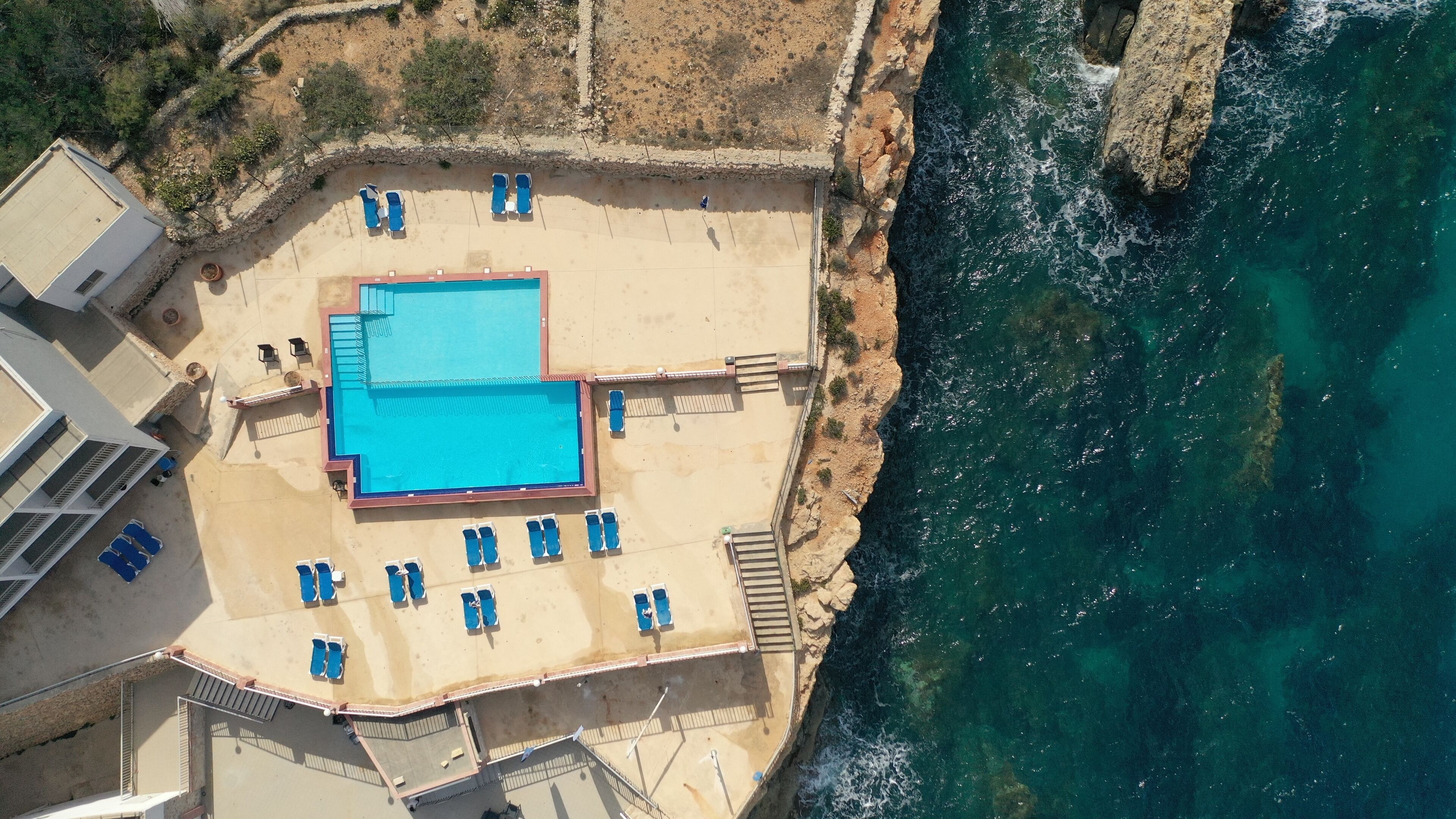 Vista aérea de un hotel frente al mar con piscina, tumbonas y un litoral rocoso.