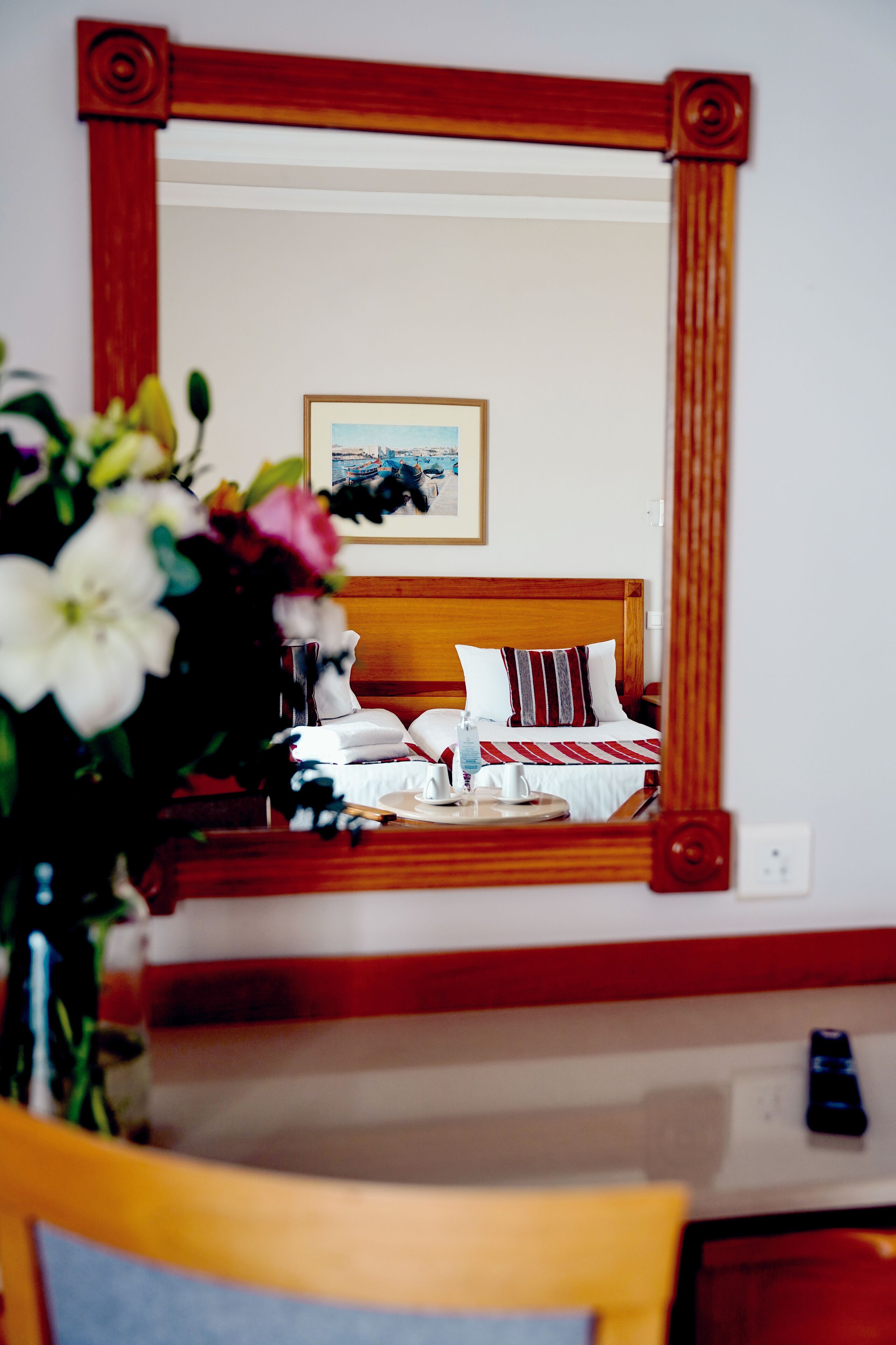 Habitación de hotel con cabecera de madera, cama con ropa de cama blanca y a rayas, un cuadro en la pared y un florero con flores sobre la mesita de noche.