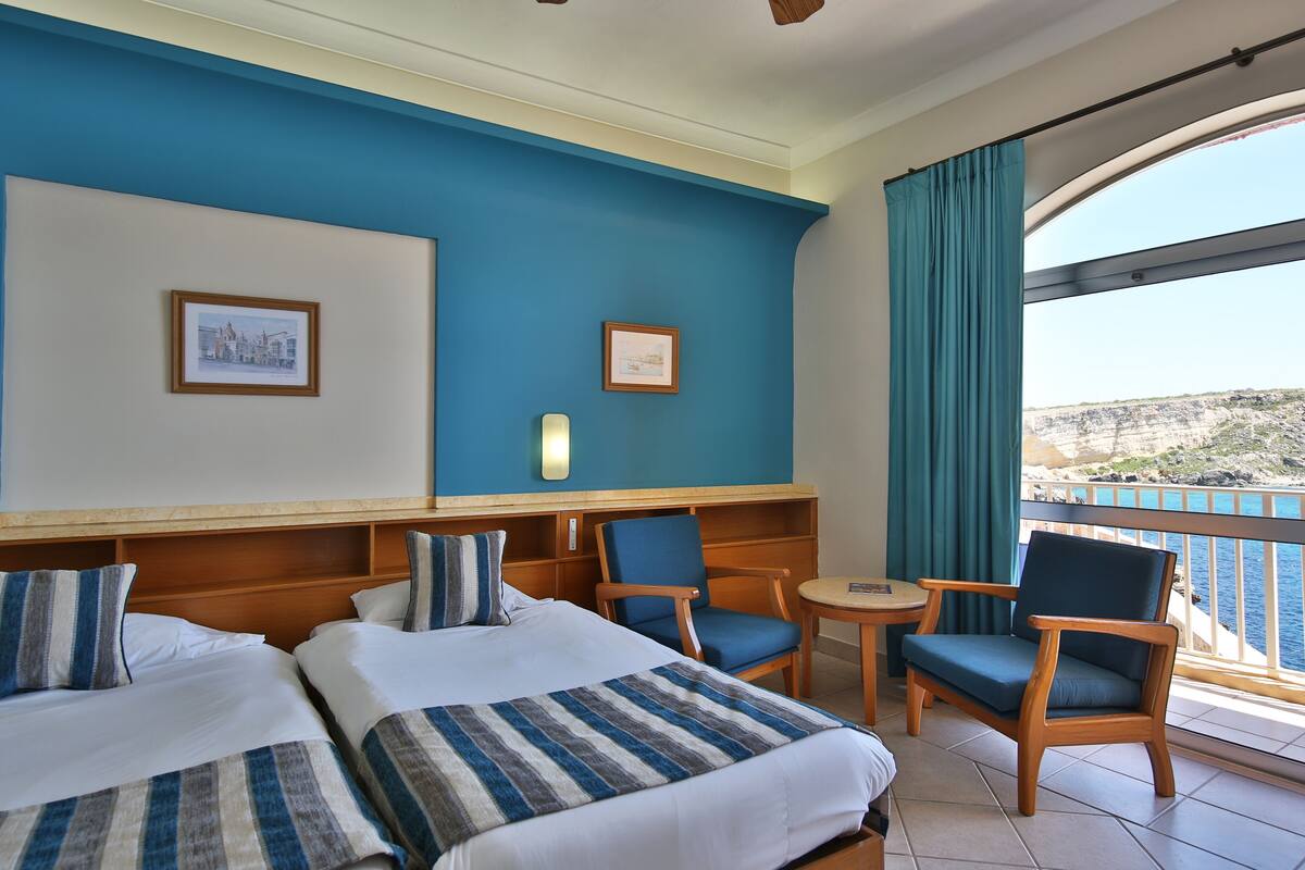 A hotel room with two beds, a seating area with chairs, a small table, and a balcony with a view of the sea.