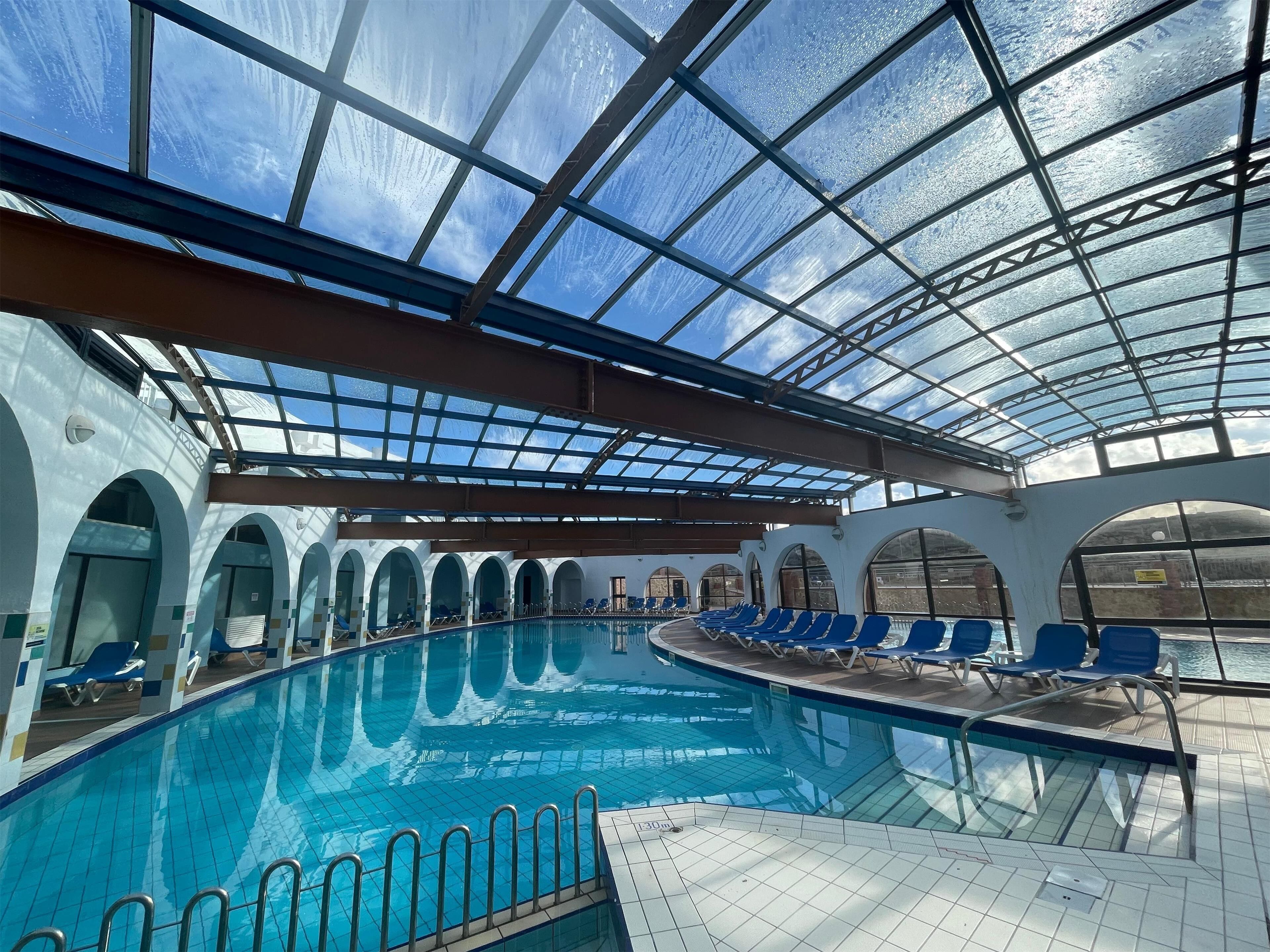 An indoor swimming pool with a glass ceiling, blue chairs around the pool, and arched doorways.