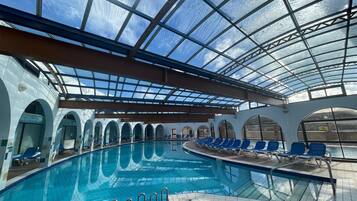 An indoor swimming pool with a glass ceiling, blue chairs around the pool, and arched doorways.
