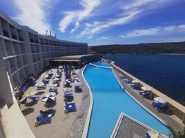 Area piscina con sedie a sdraio e ombrelloni, una grande piscina e un edificio con numerosi balconi.