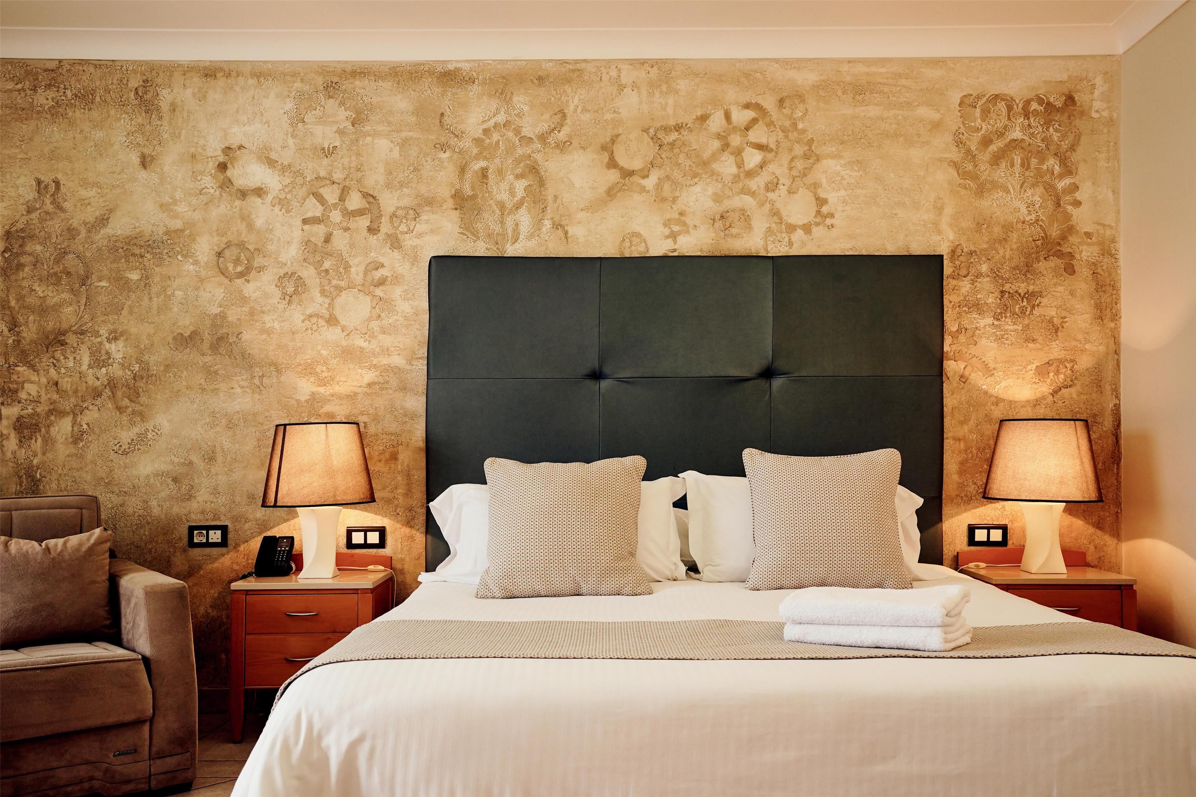 A hotel room with a large bed, two bedside tables with lamps, a chair, and a textured wall.