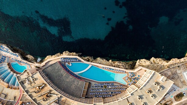 Luchtbeeld van een resort met meerdere zwembaden, ligbedden en een rotsachtige kustlijn.