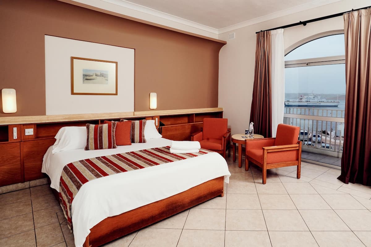 A hotel room with a large bed, a seating area with a table, a view of a ship through the window, and a framed picture on the wall.