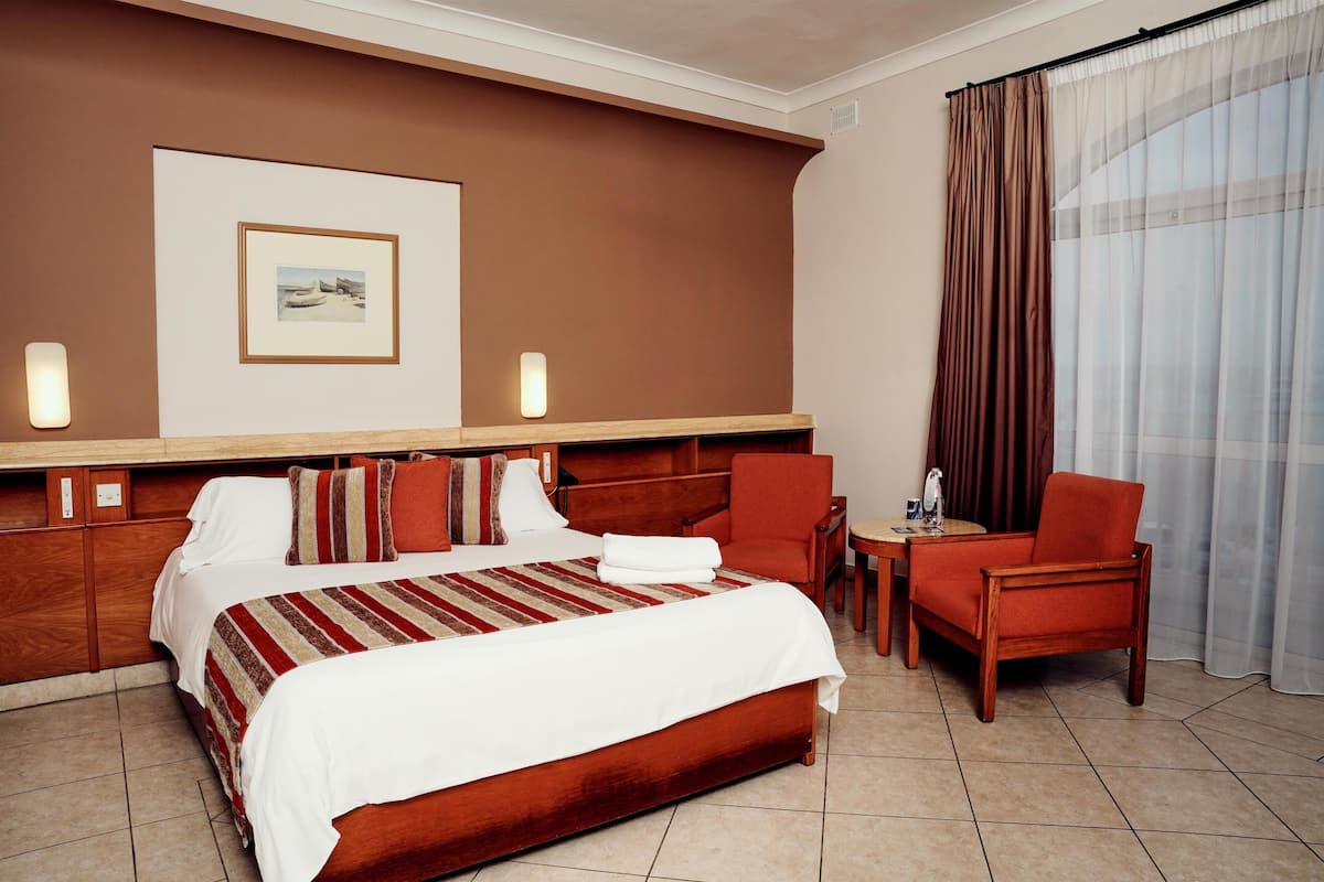A hotel room with a large bed, a wooden headboard, a small table, a chair, and a window with curtains.