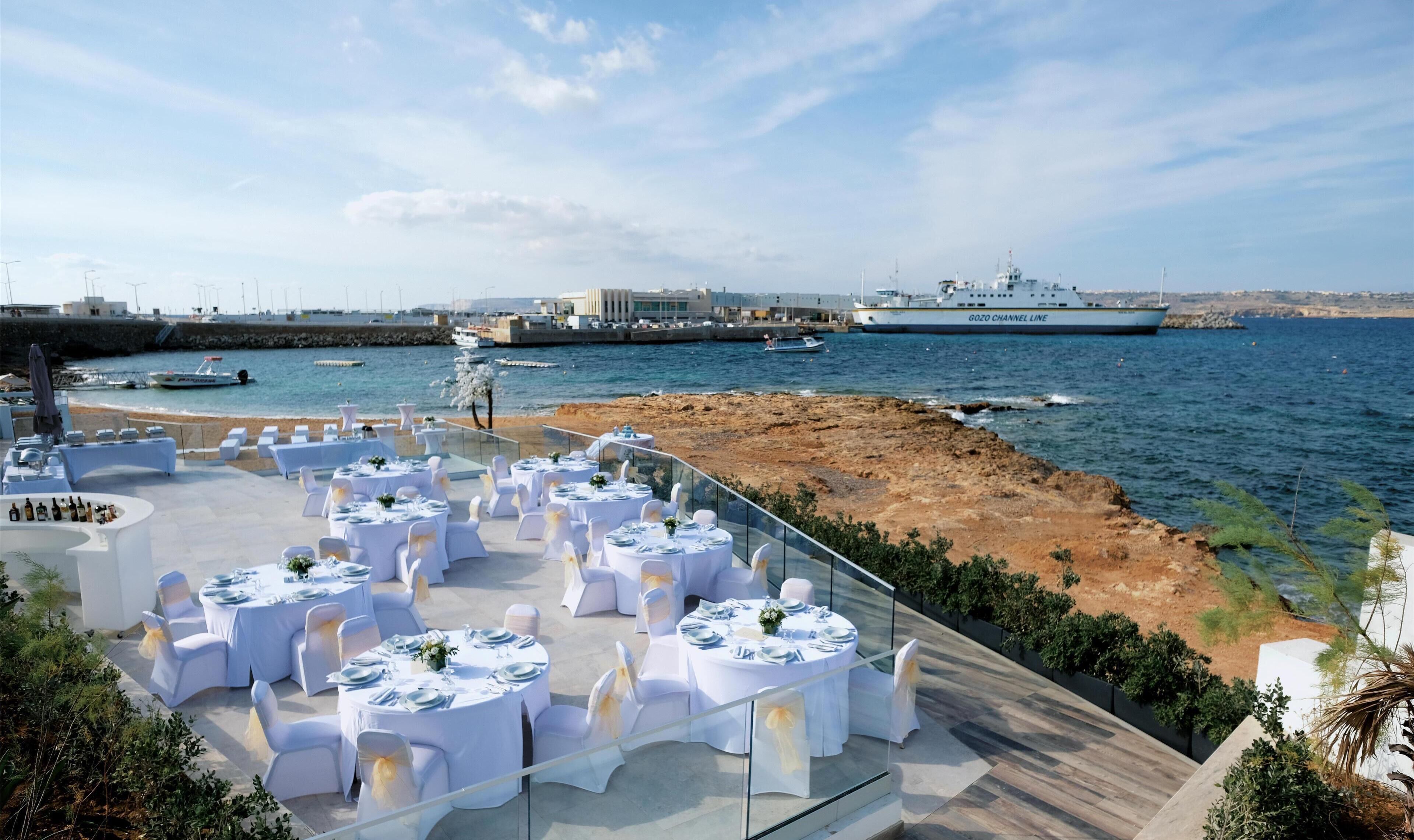 An outdoor dining area with round tables set for a wedding, overlooking a rocky coastline and the sea.