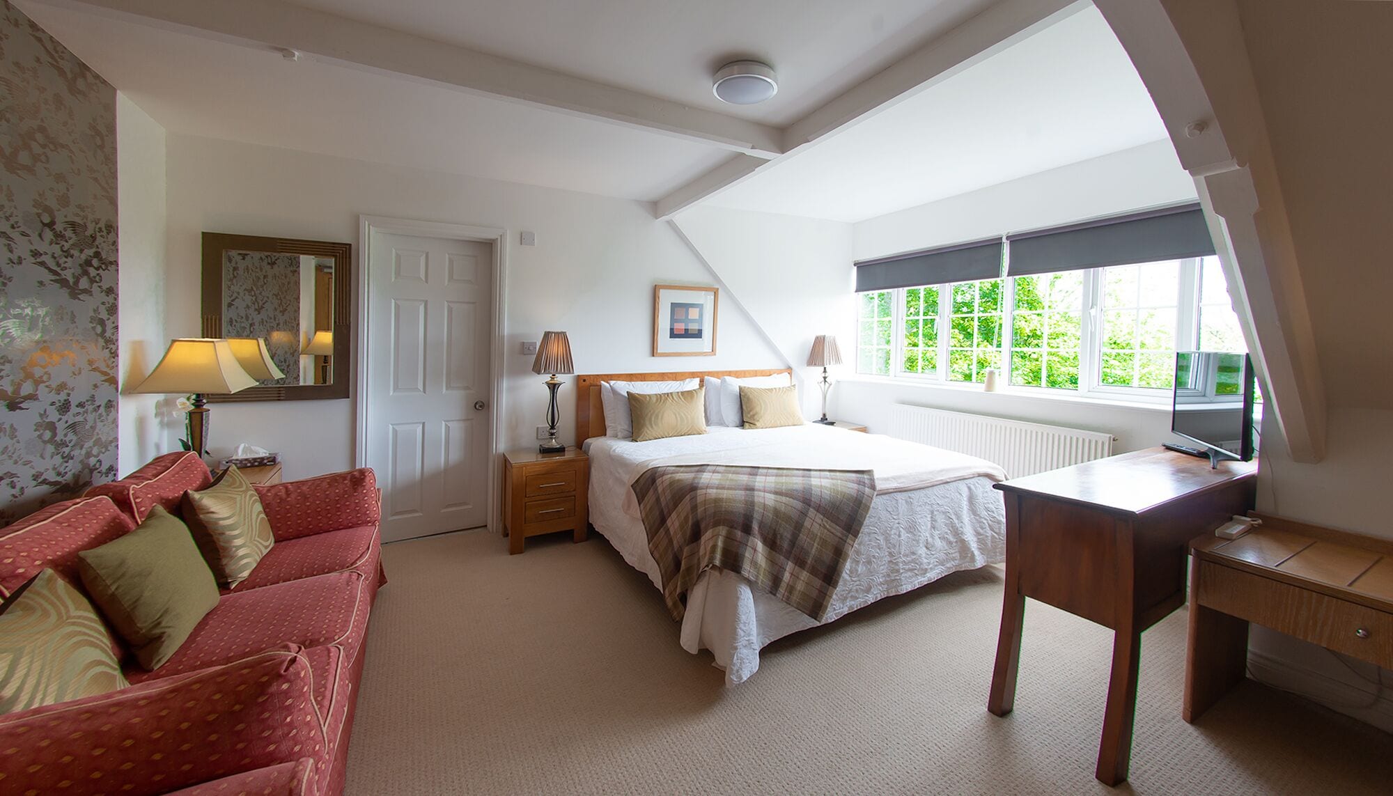 A bedroom with a bed, a desk, a chair, a red sofa, a window with a view of greenery, and a door.